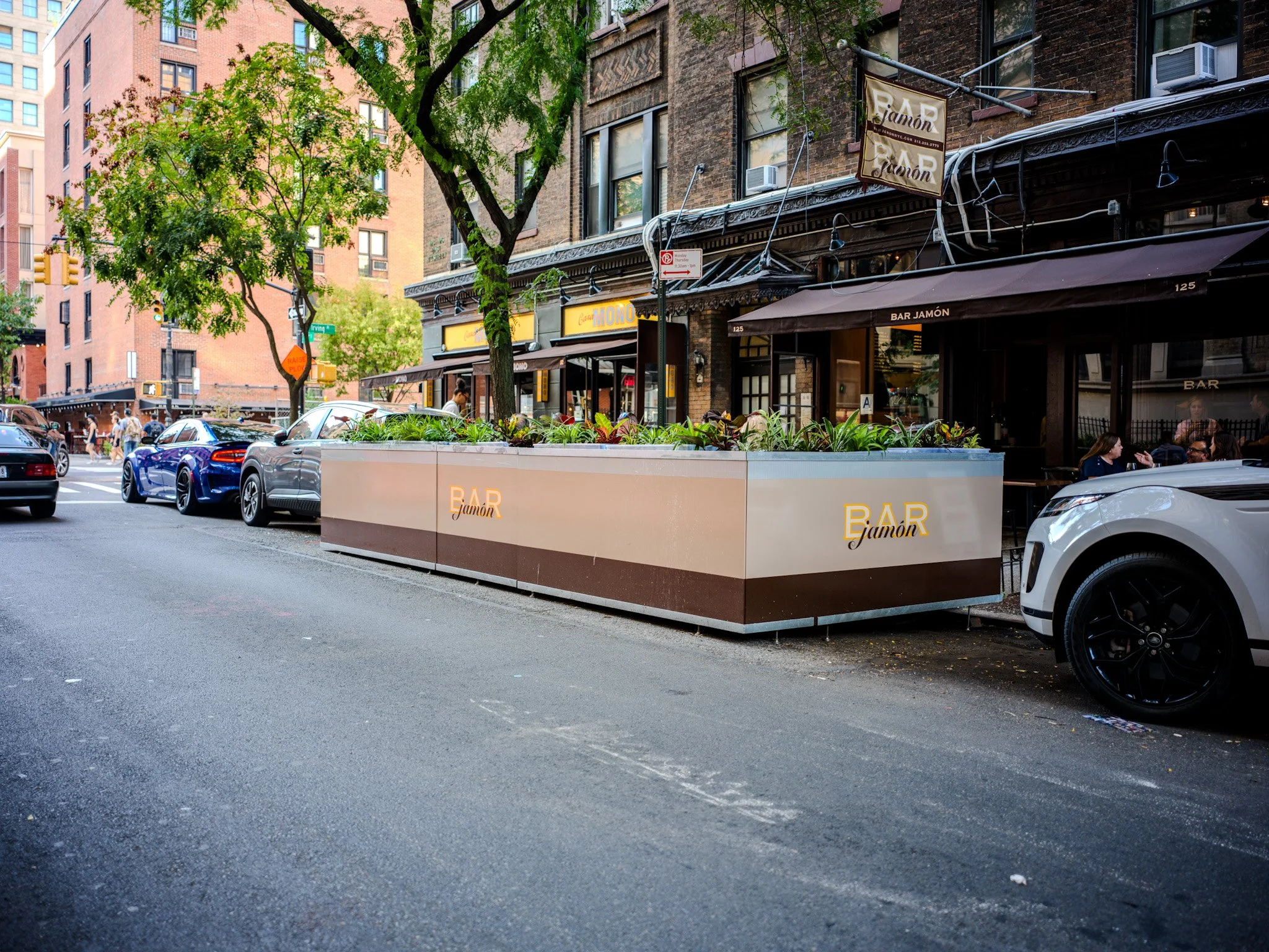 City street scene with parked cars, a sidewalk café, and a large planter with greenery in front of a brick building, featuring signs for a bar called 'Bar Jamón'.