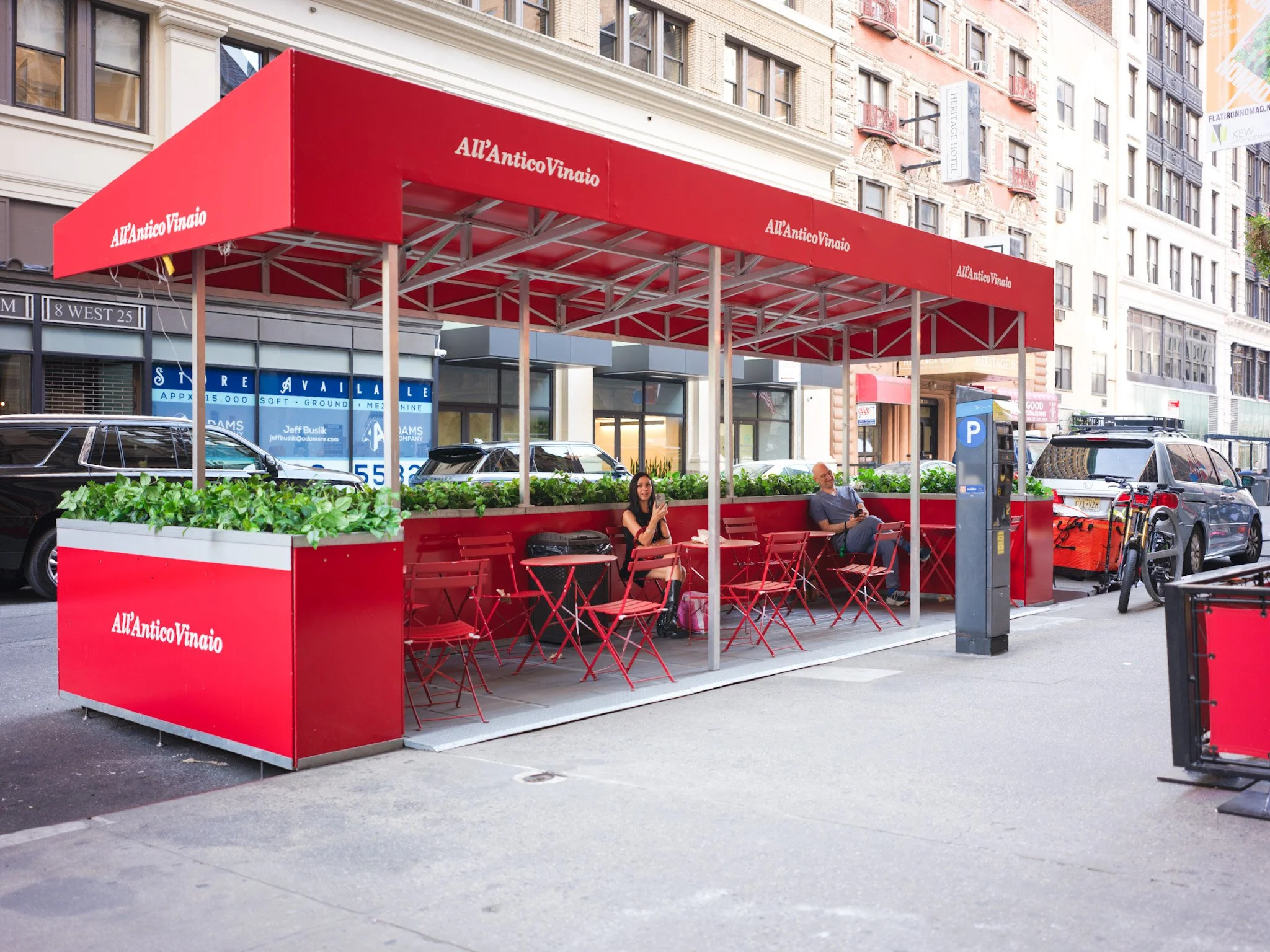 Red outdoor seating area with tables and chairs, marked with 'All'AnticoVinaio', on a city sidewalk. Two people are sitting and using their phones, behind the enclosure is parked cars and buildings.