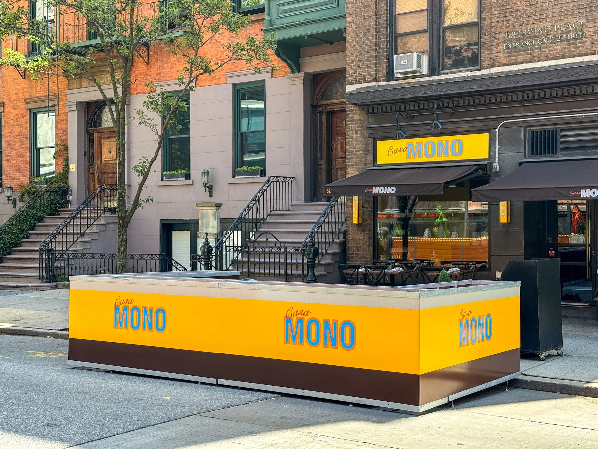 Yellow and brown outdoor food stall with the sign Casa Mono, parked on the sidewalk in a city street in front of brownstone buildings with stoops and trees.