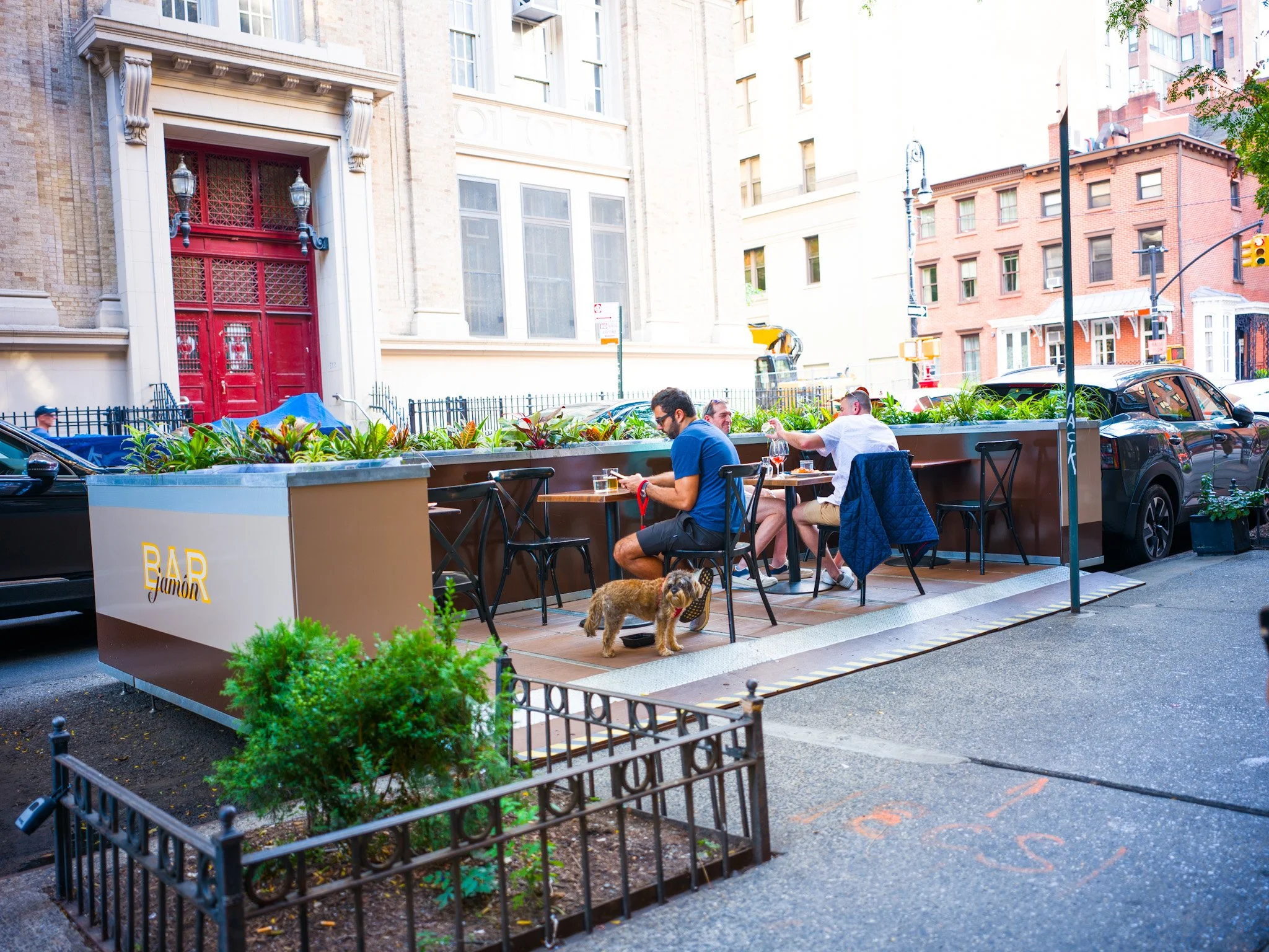 City sidewalk cafe with three men sitting at tables, one man reading, and a dog standing on the ground nearby. Surrounding buildings and parked cars are visible.