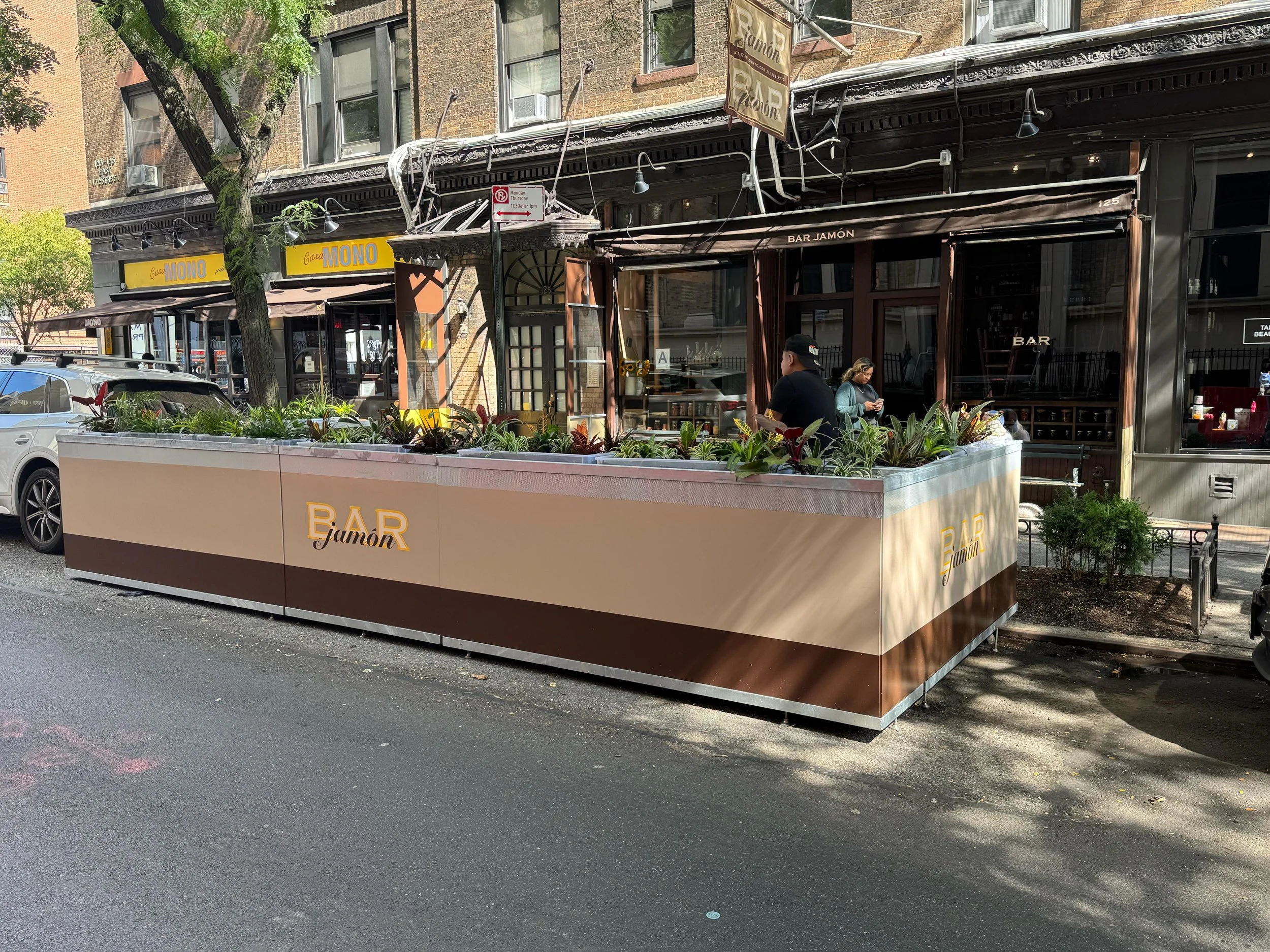 Street view of a restaurant named Bar Jamón with outdoor seating, plants in planters, pedestrians, and parked cars along the street.