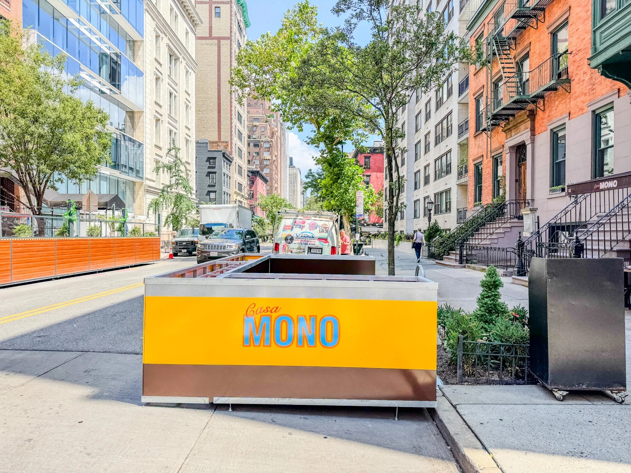 Street view with a yellow and brown trash container labeled 'Casa MONO' in the foreground. Tall buildings on either side, trees lining the sidewalk, parked cars, and a few pedestrians. Bright, sunny day.