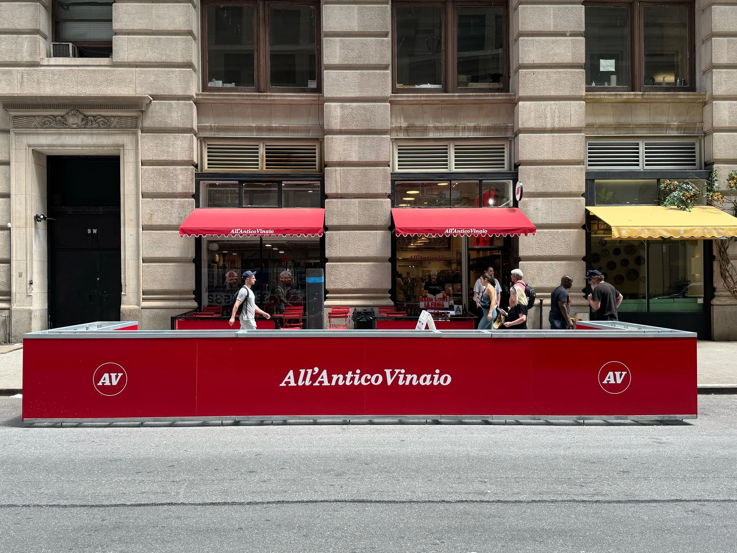 Street scene with a red outdoor seating area in front of a restaurant named 'All'Antico Vinaio'. Several pedestrians walk by; some are entering or exiting the restaurant. The building has a stone facade with large windows and awnings.