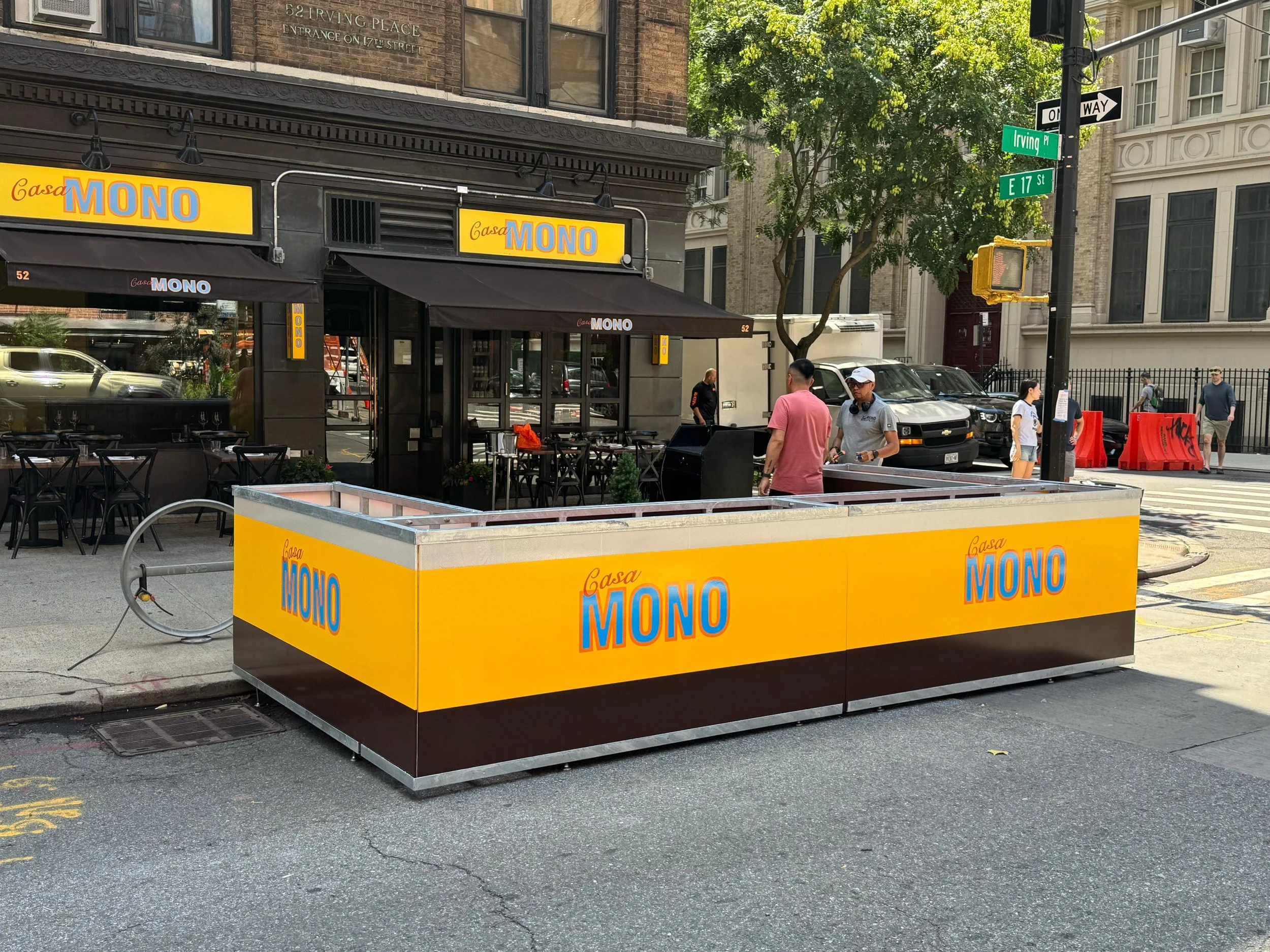Outdoor seating area in front of a restaurant called Casa Mono, with yellow signage, black awnings, and black chairs. A temporary bright yellow and black structure with the same restaurant name is on the sidewalk. Several people are walking or standi