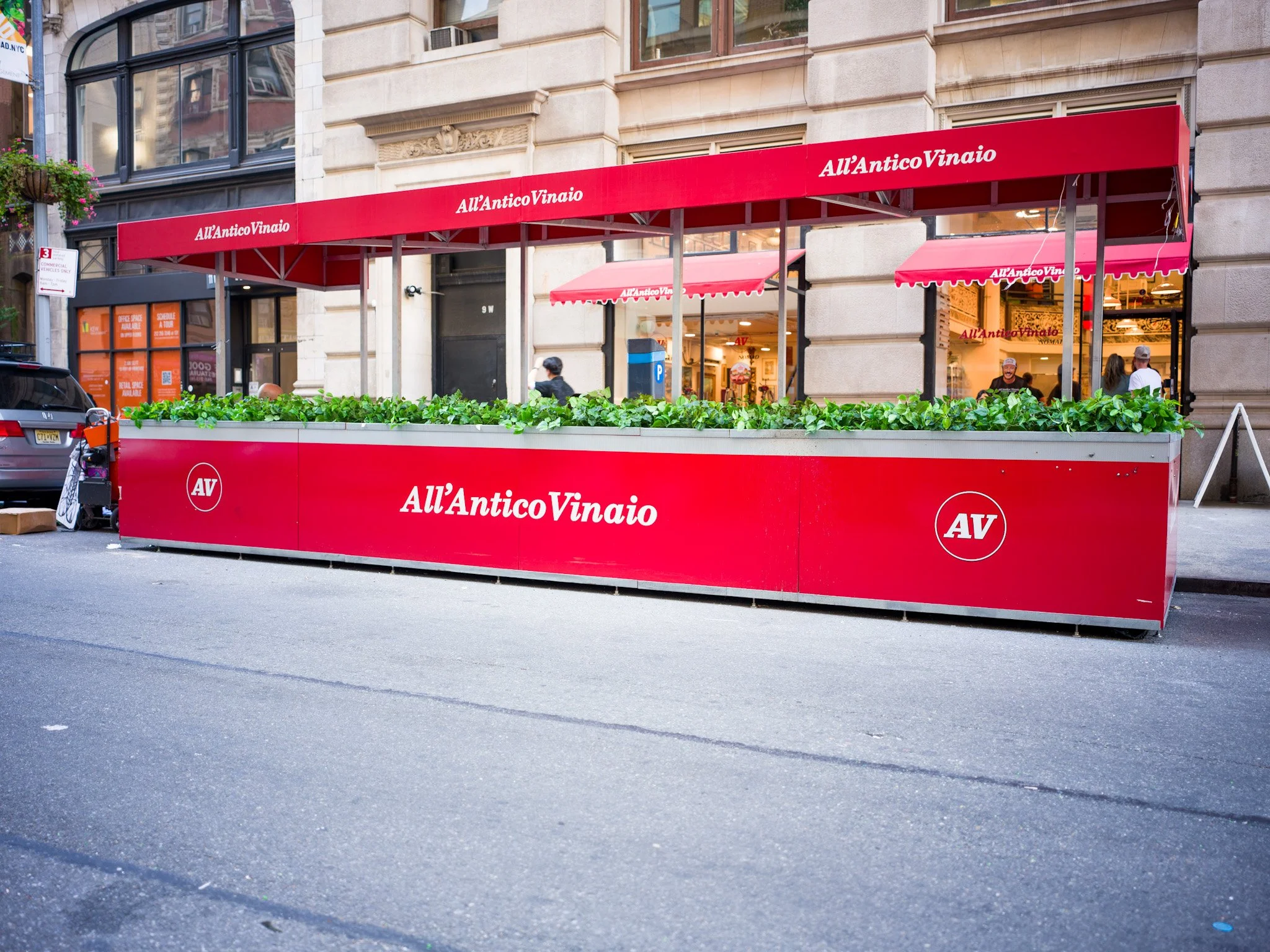 Red outdoor patio fencing with plants and signage for All’Antico Vinaio, a restaurant, with a building facade and people inside visible in the background.