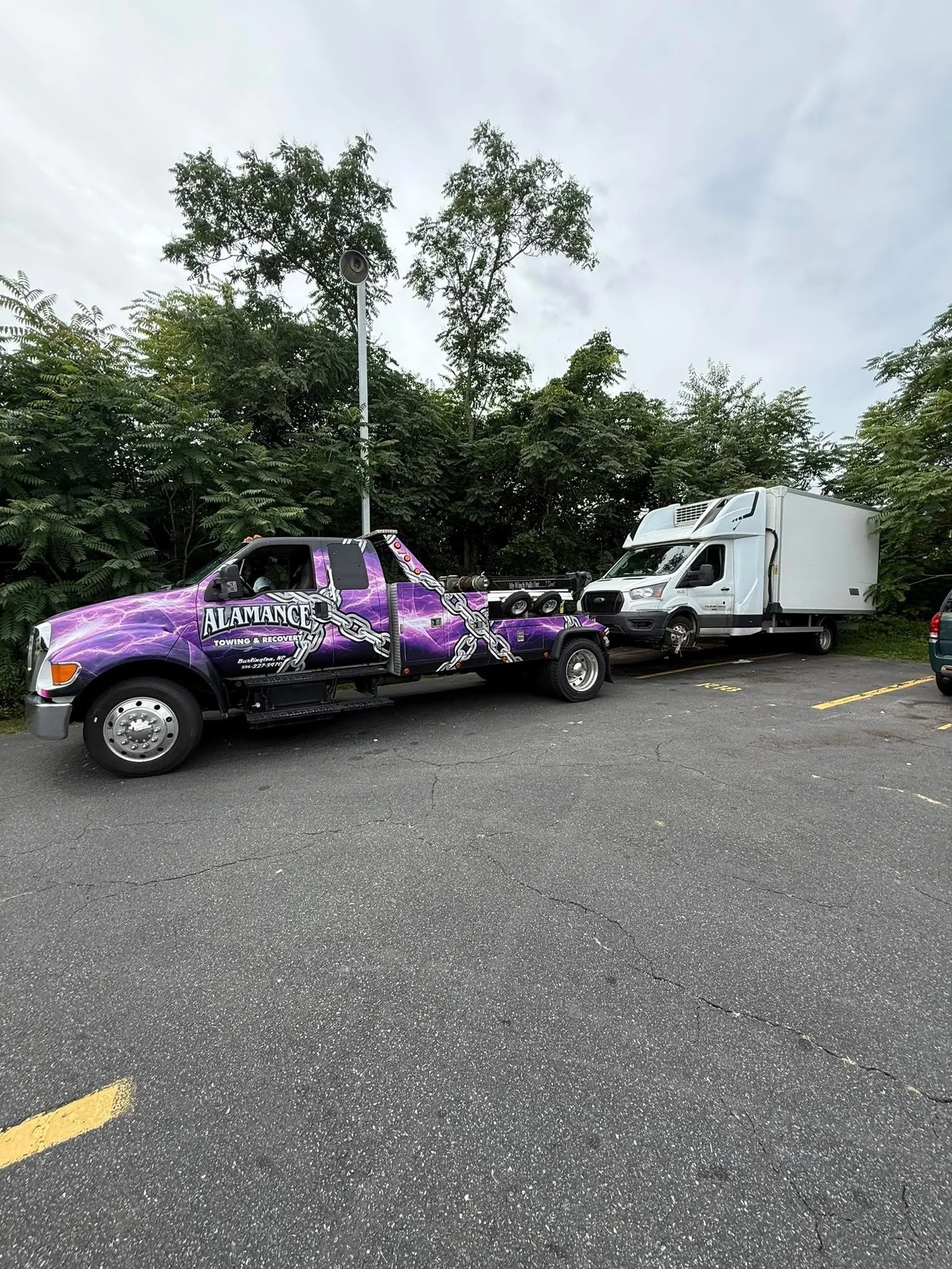 A tow truck with a purple lightning design labeled 'ALAMANDE Towing & Recovery' is towing a white delivery truck in a parking lot surrounded by trees.