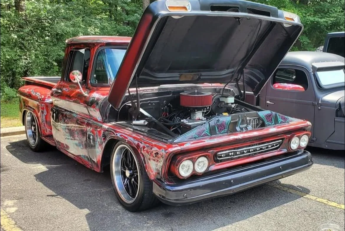 A vintage red Chevrolet pickup truck with a custom paint job and open hood revealing a modified engine, parked in a lot next to another classic gray vehicle, surrounded by greenery.