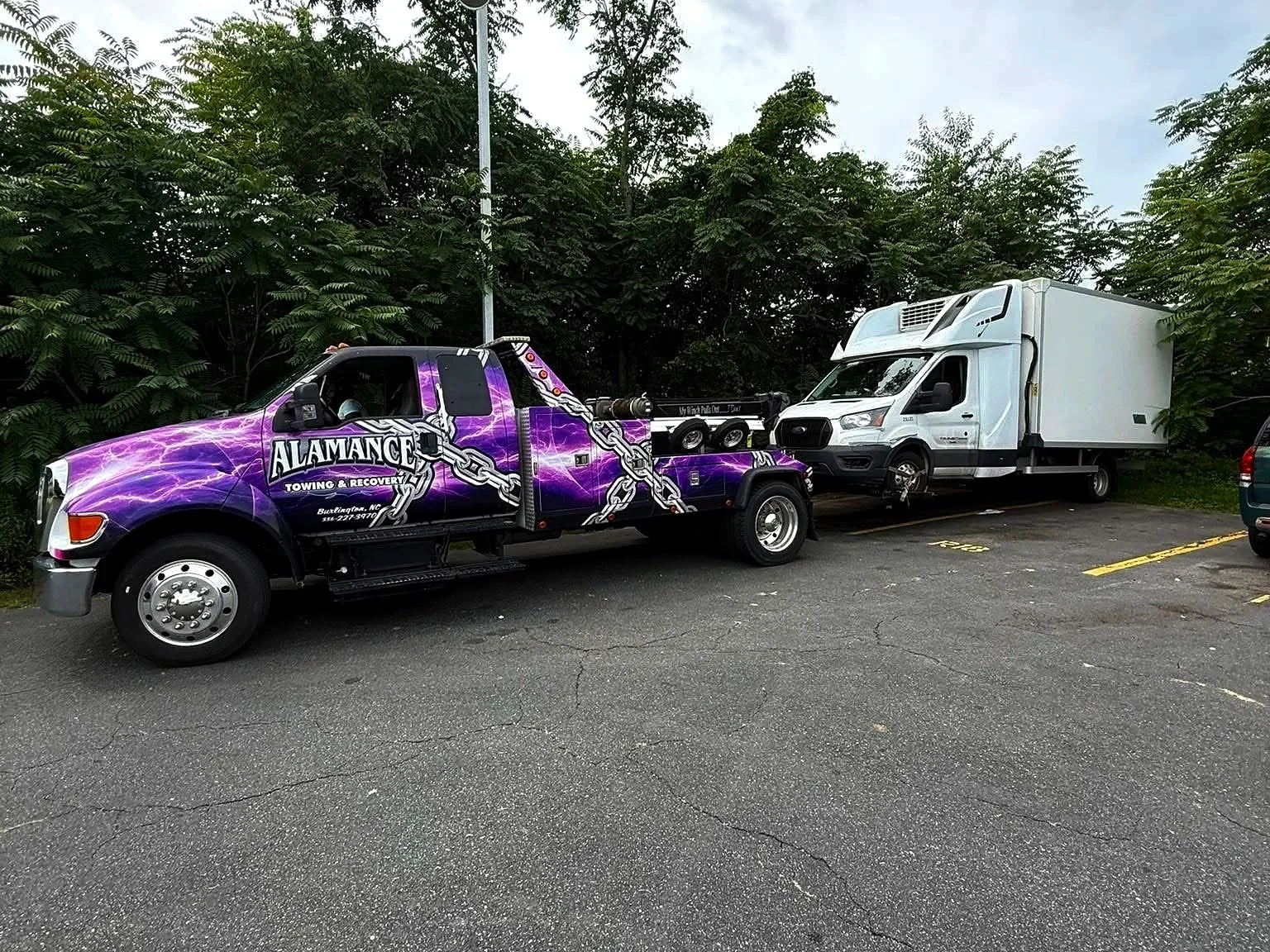 A tow truck with a purple lightning design labeled 'ALAMANDE Towing & Recovery' is towing a white delivery truck in a parking lot surrounded by trees.