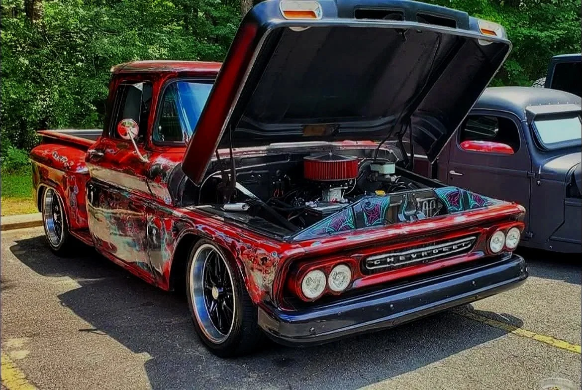 A vintage red Chevrolet pickup truck with a custom paint job and open hood revealing a modified engine, parked in a lot next to another classic gray vehicle, surrounded by greenery.