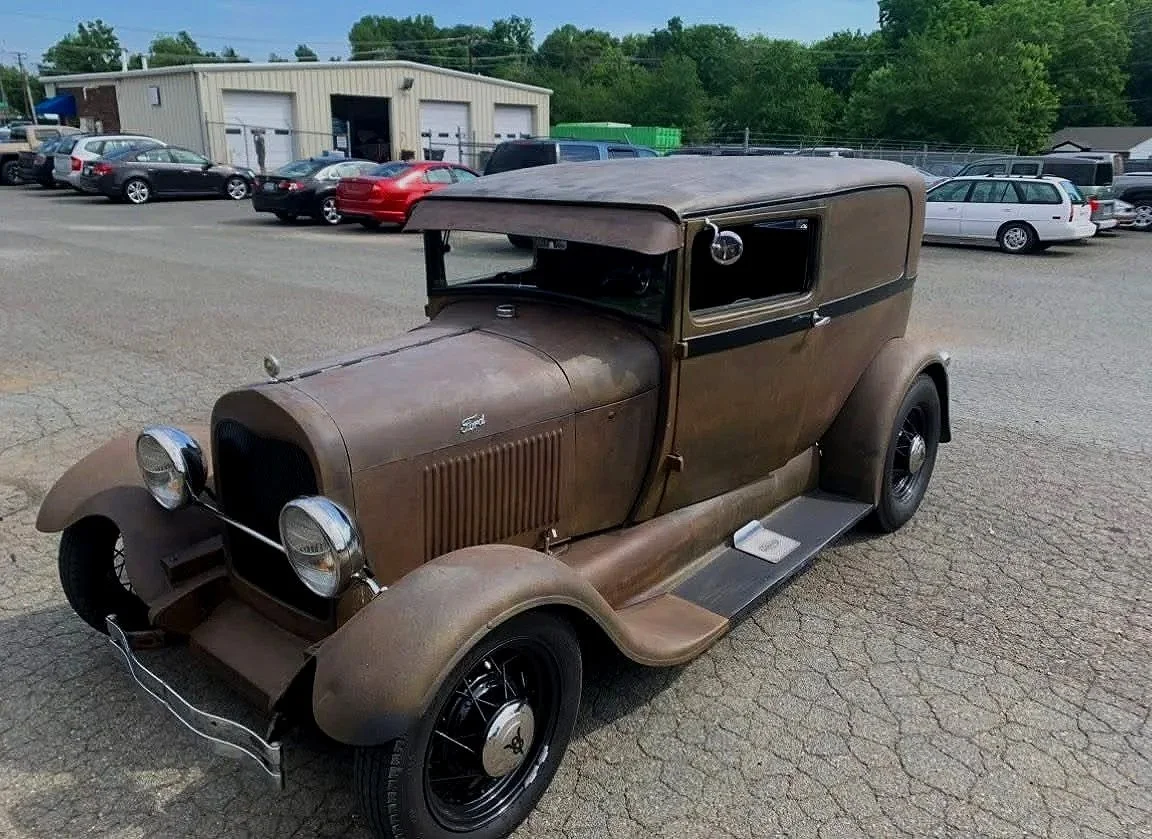 Old, rusty, vintage car parked in a lot with other vehicles and a building in the background.