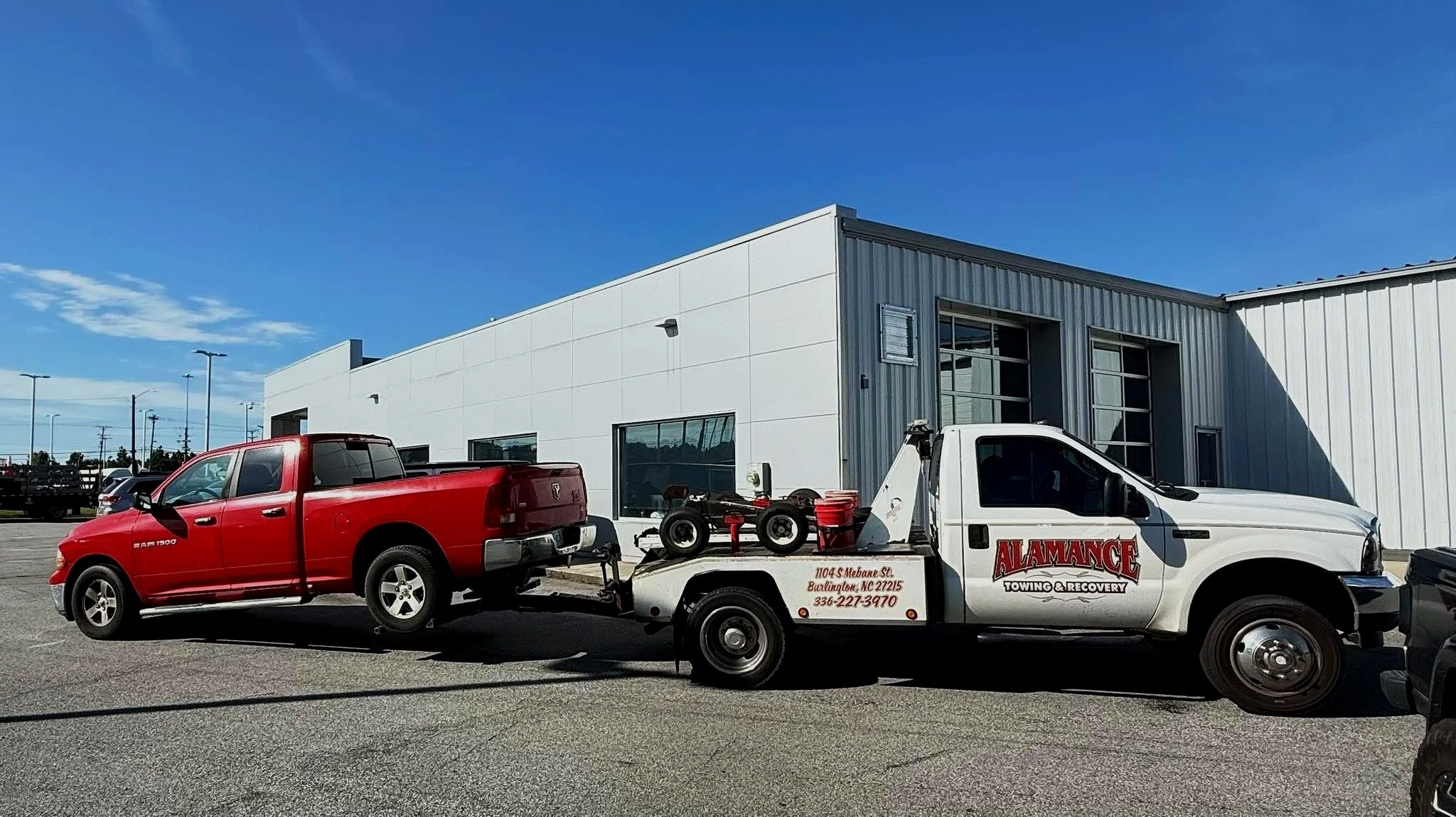 A white tow truck transporting a red pickup truck parked outside a modern commercial building under a blue sky.