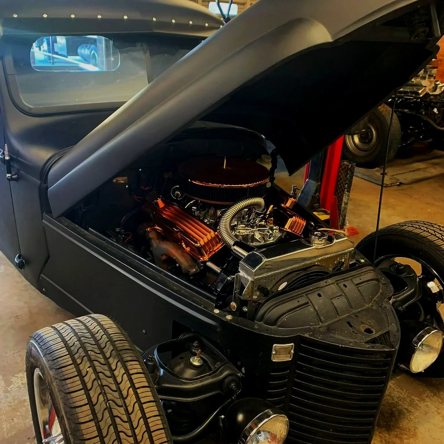 A black vintage hot rod car with its hood open, showing the engine with copper accents, in a garage with other vehicle parts and equipment in the background.