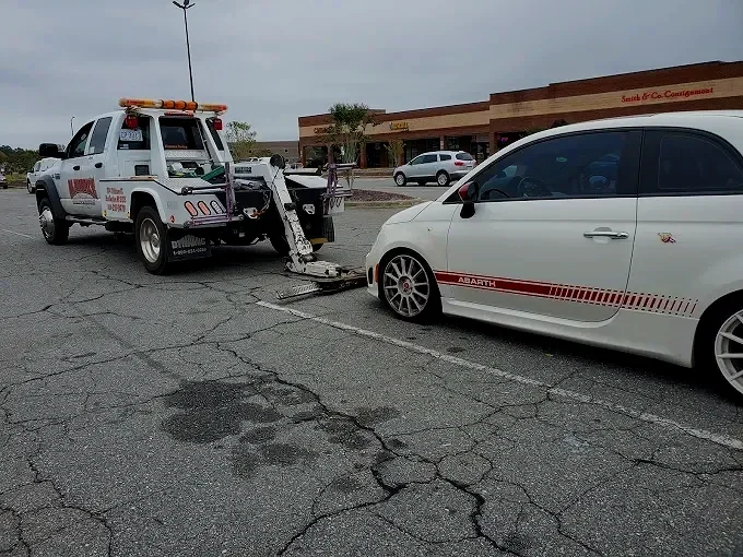 A white tow truck with a flatbed and orange safety lights is towing a white compact car with red stripe graphics in a parking lot of a shopping center. The parking lot has cracked pavement, and there are a few other cars and a store with a sign that 