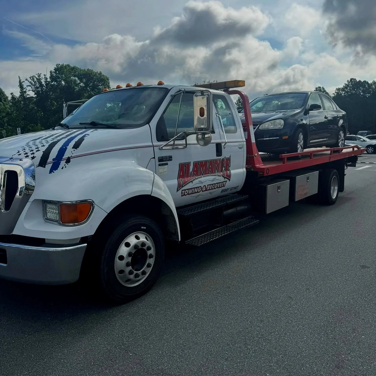 Tow truck carrying a black sedan in a parking lot with a cloudy sky