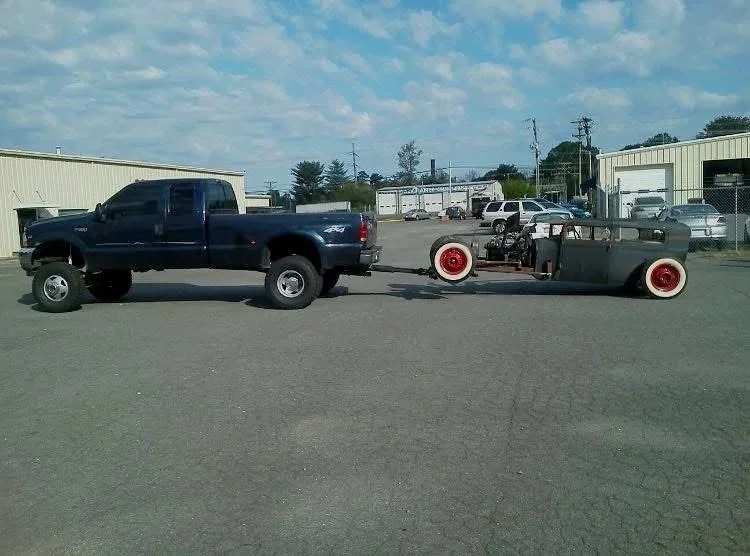 A large black pickup truck towing a vintage race car with exposed engine and red wheels in an open lot with industrial buildings and vehicles in the background.