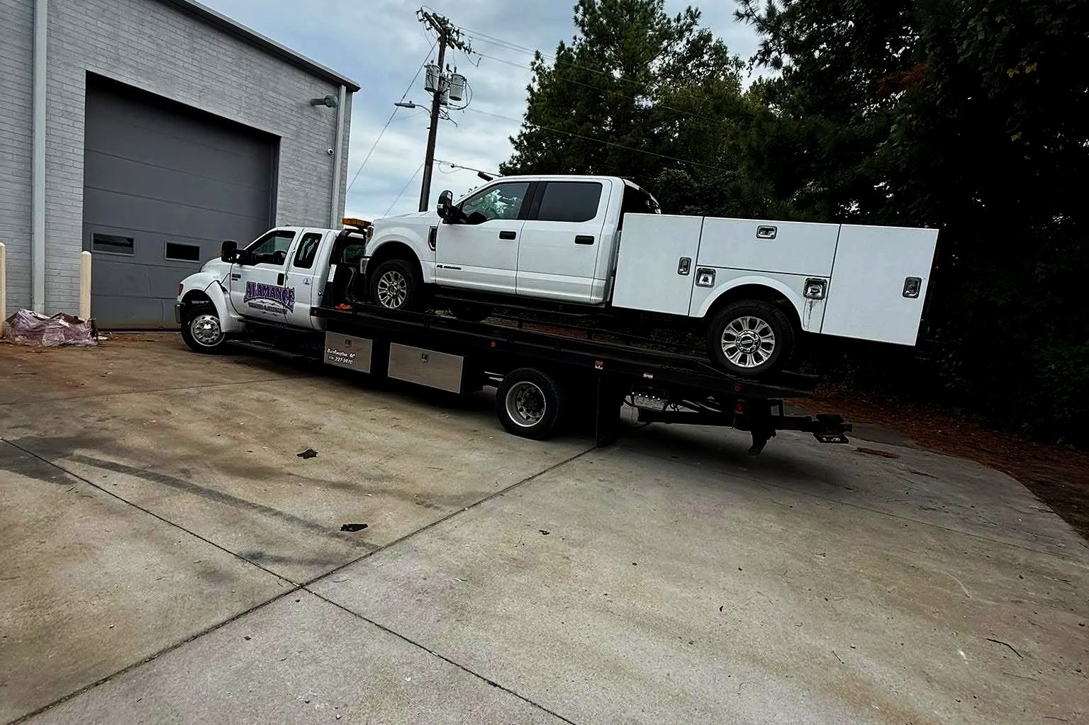 A tow truck transporting a white pickup truck parked outside a building with a large garage door, overcast sky, and trees in the background.