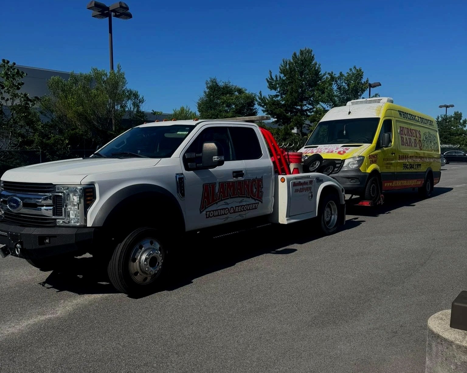 Tow truck from Alamance Towing & Recovery towing a yellow vehicle with advertising for a farm and bar, parked in a lot under a clear blue sky.