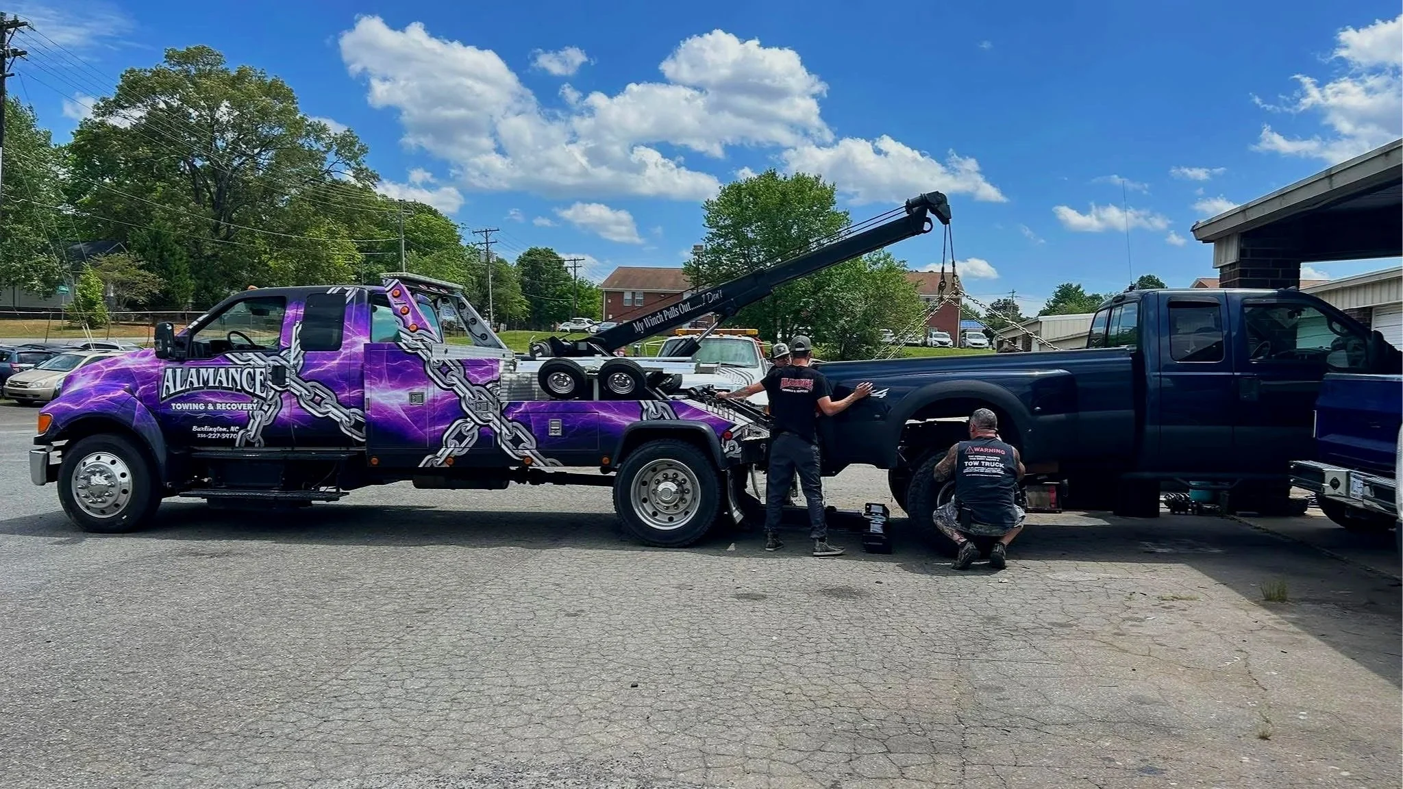 A tow truck painted purple with chains logos towing a black pickup truck in a parking lot on a sunny day, with four workers preparing the vehicle.