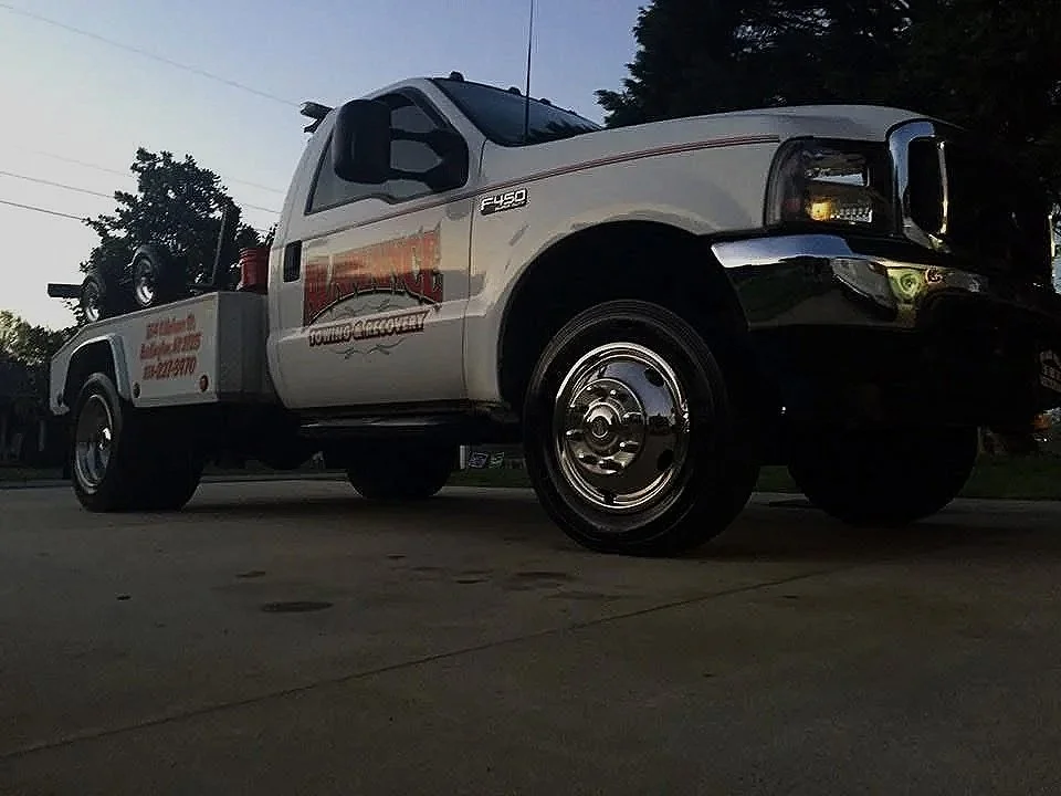 A white tow truck with a logo on the door, parked on a paved surface during dusk. A spare tire is mounted on the truck bed, and the vehicle has chrome hubcaps.