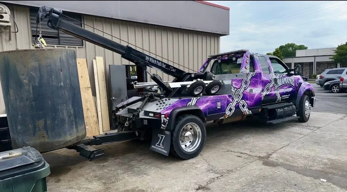 A tow truck with a purple and white chain design parked in a lot near a building, with a large black object secured at the back.