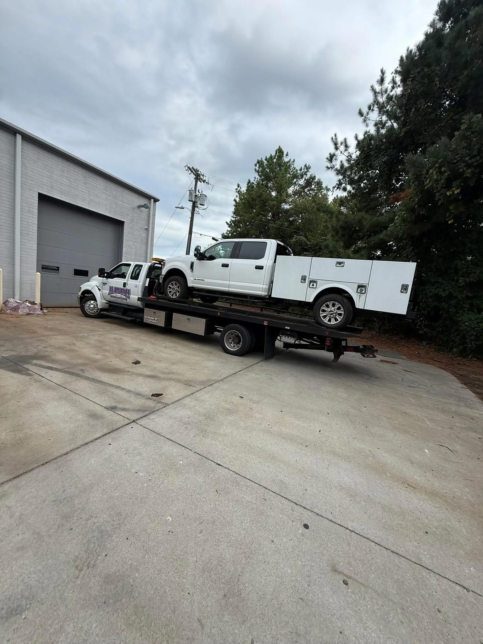 A tow truck transporting a white pickup truck parked outside a building with a large garage door, overcast sky, and trees in the background.