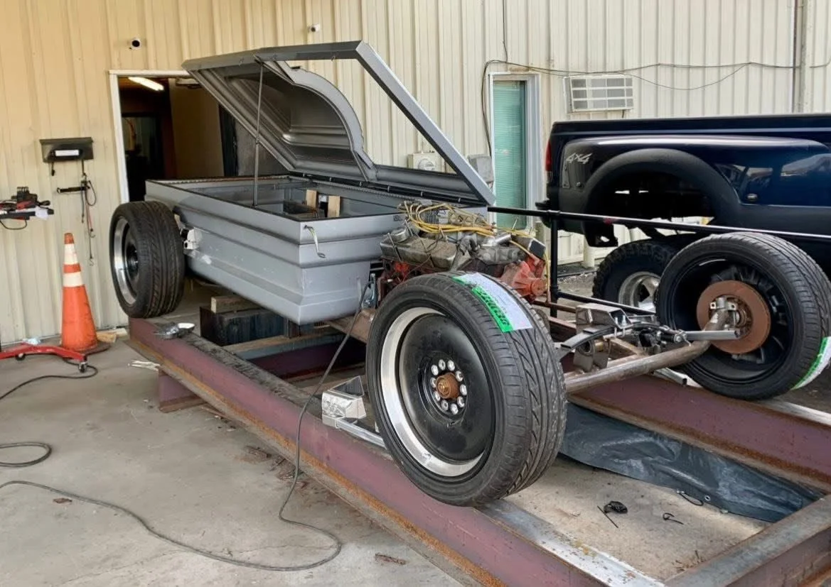 A partially assembled race car with a metal body, large tires, and exposed engine components on a metal platform in a workshop.