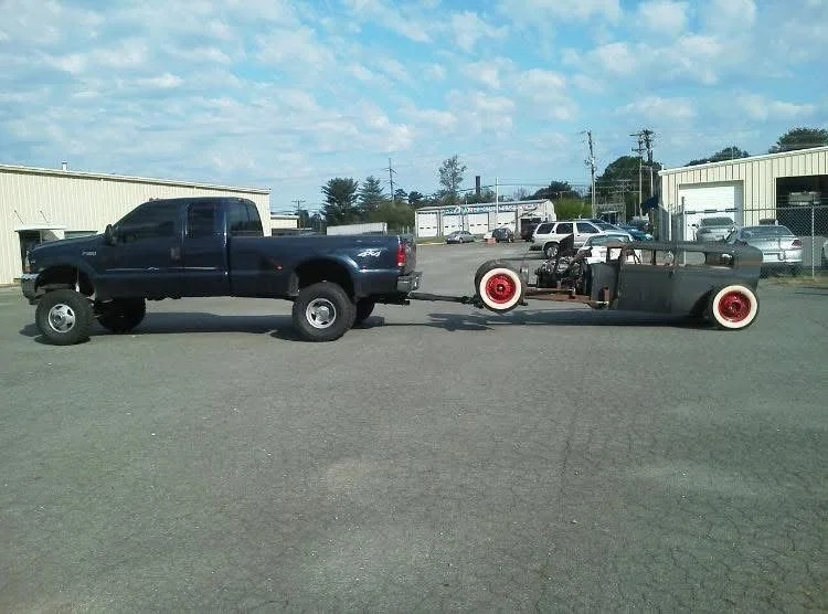 A large black pickup truck towing a vintage race car with exposed engine and red wheels in an open lot with industrial buildings and vehicles in the background.