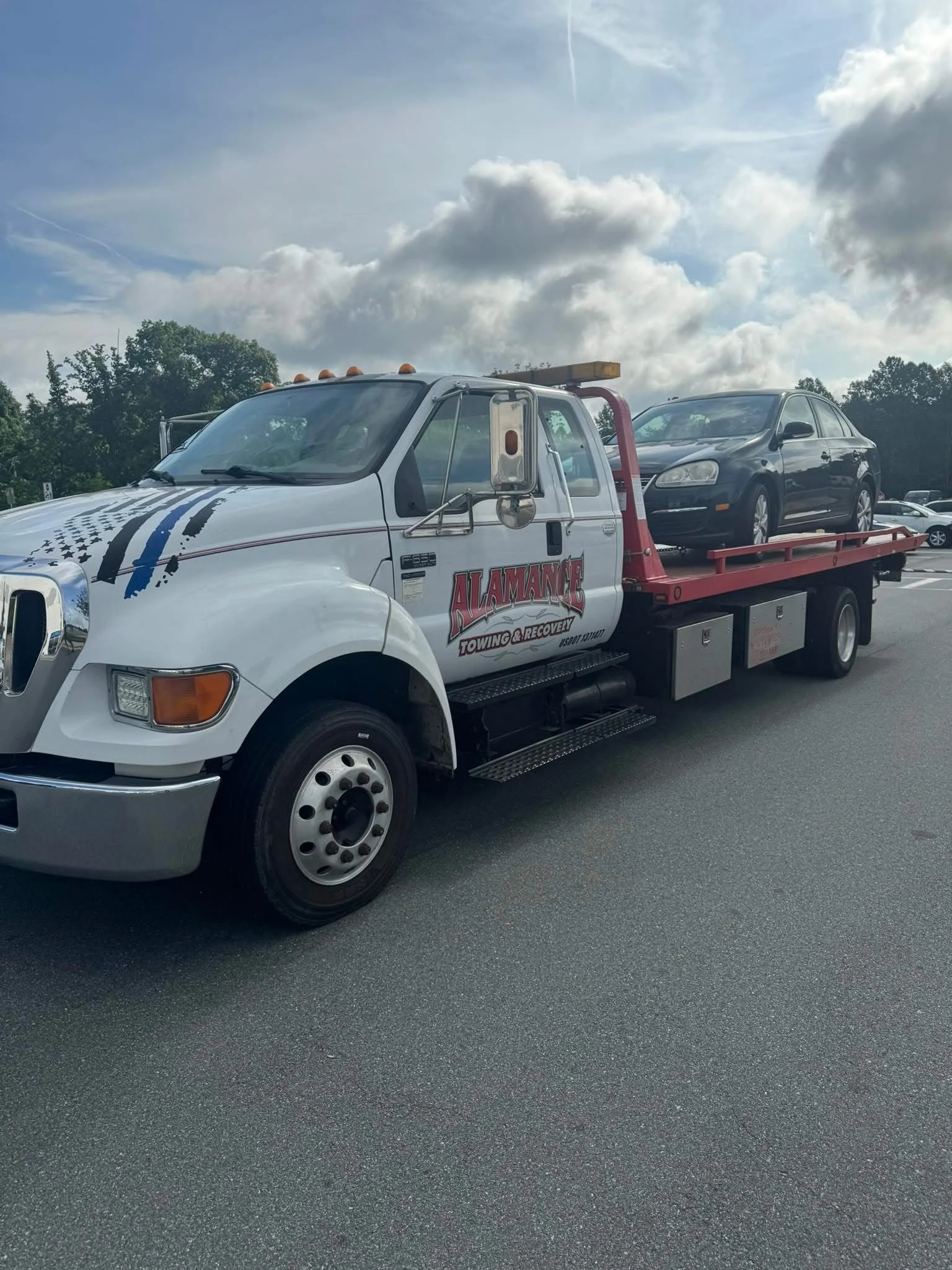 White tow truck labeled "Alamance Towing & Recovery" carrying a black sedan in a parking lot under a cloudy sky.