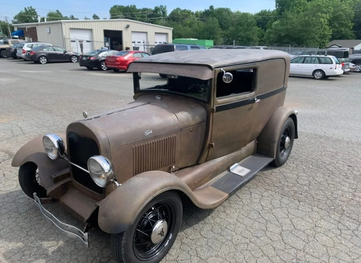 Old, rusty, vintage car parked in a lot with other vehicles and a building in the background.