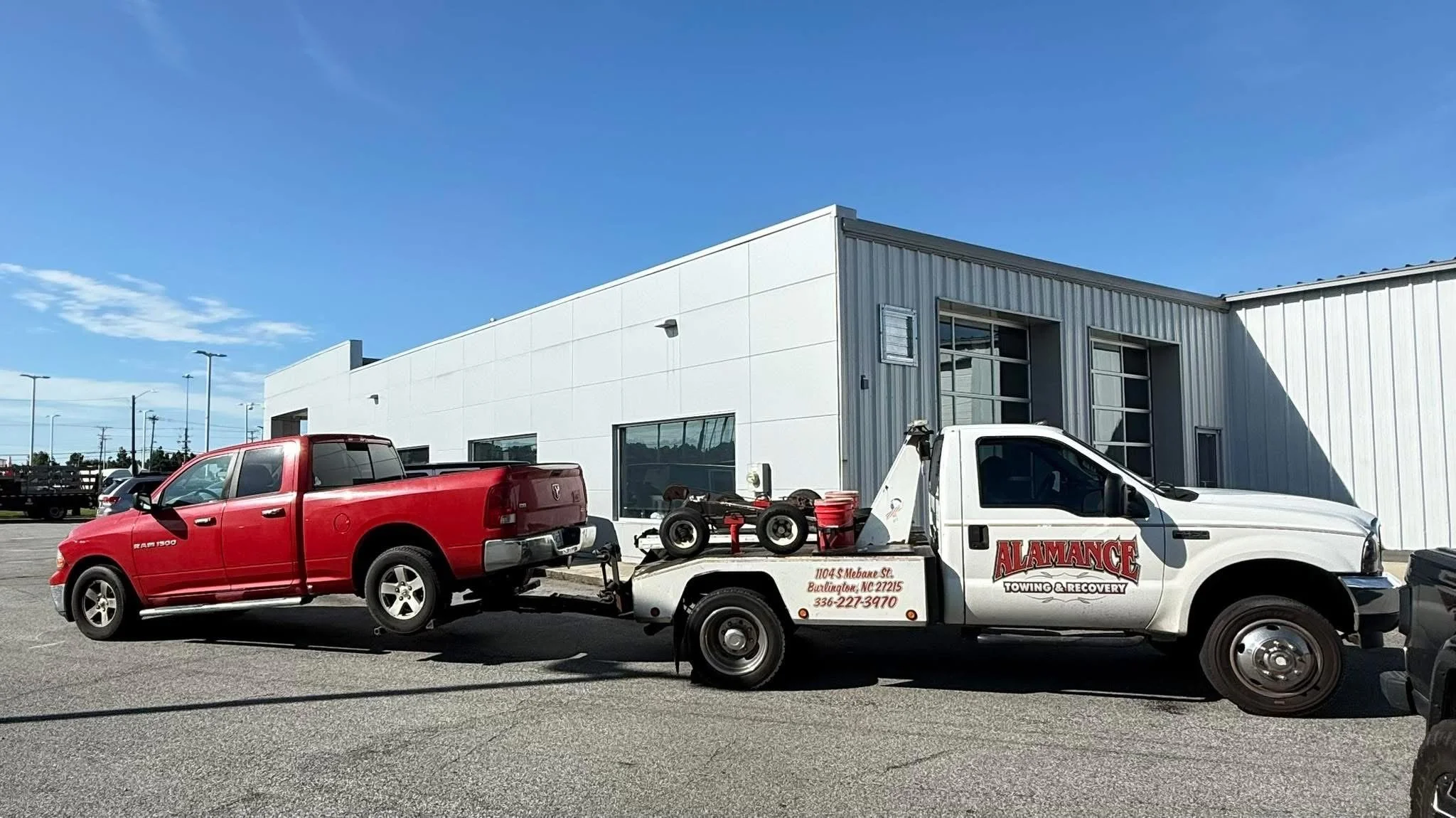 A white tow truck transporting a red pickup truck parked outside a modern commercial building under a blue sky.