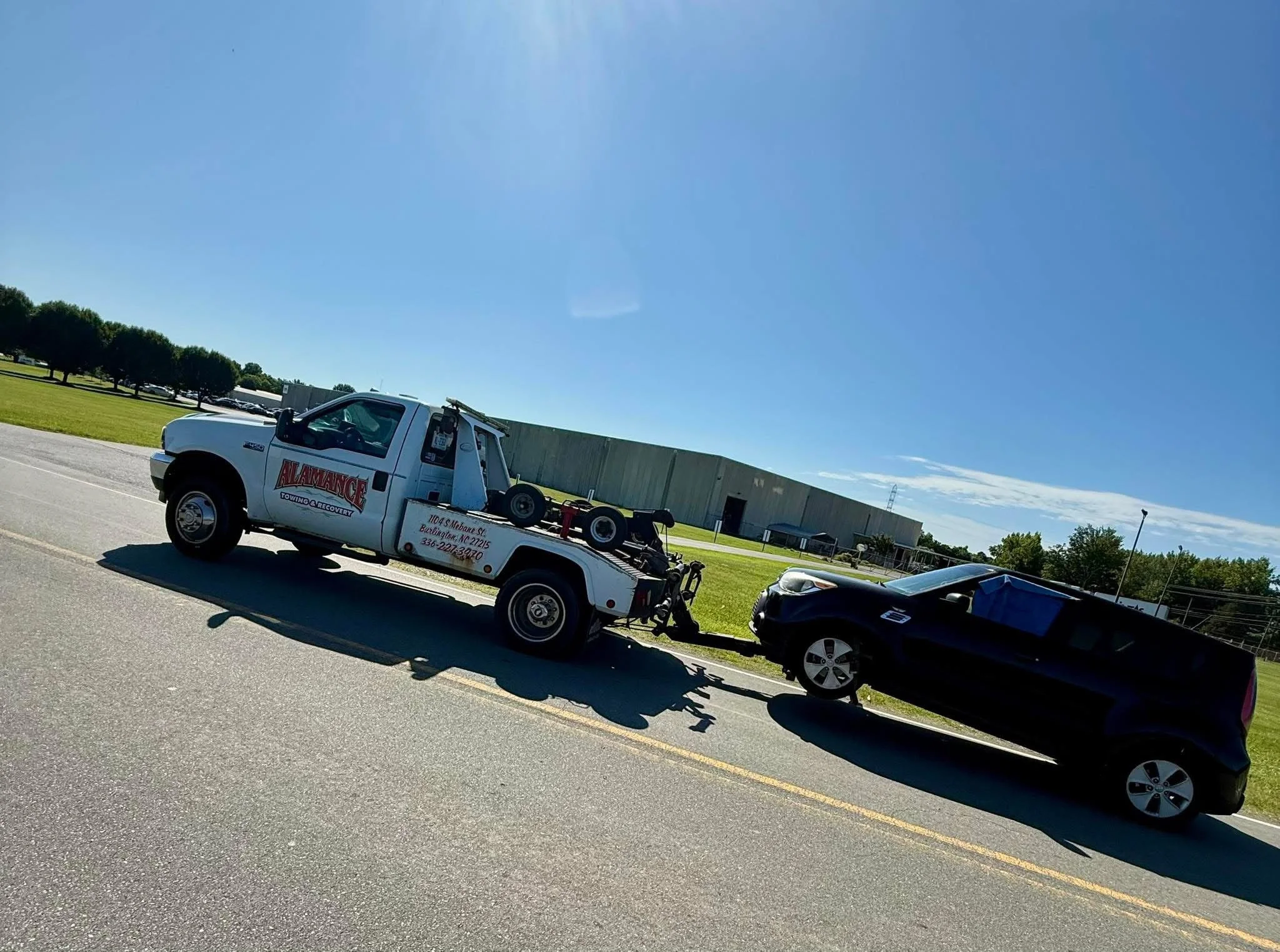 A tow truck is towing a black car on a sunny day in a suburban or industrial area.
