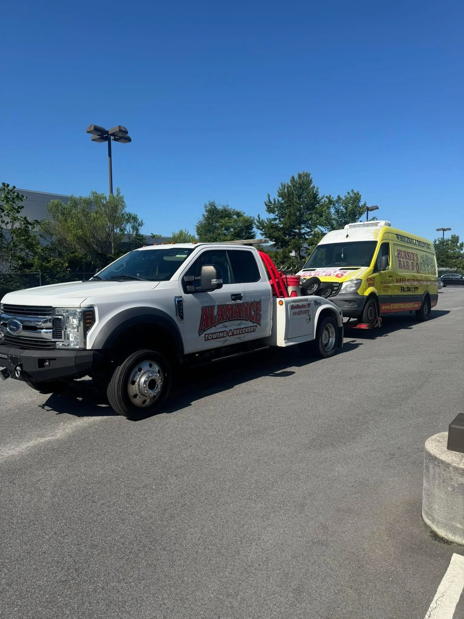Tow truck from Alamance Towing & Recovery towing a yellow vehicle with advertising for a farm and bar, parked in a lot under a clear blue sky.