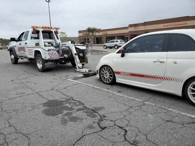 A white tow truck with a flatbed and orange safety lights is towing a white compact car with red stripe graphics in a parking lot of a shopping center. The parking lot has cracked pavement, and there are a few other cars and a store with a sign that 