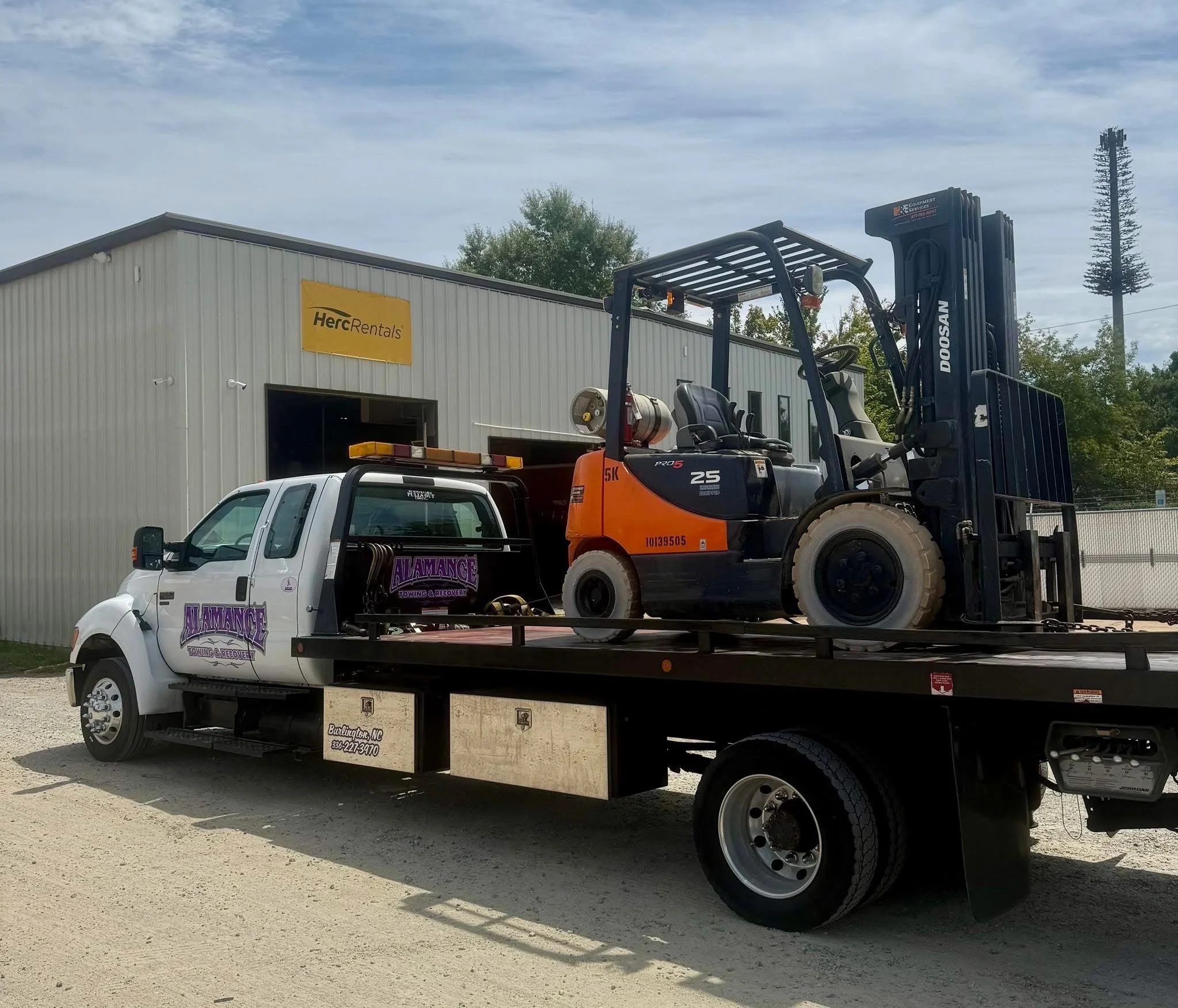 A flatbed tow truck carrying a small forklift in front of a building with a 'Herc Rentals' sign.
