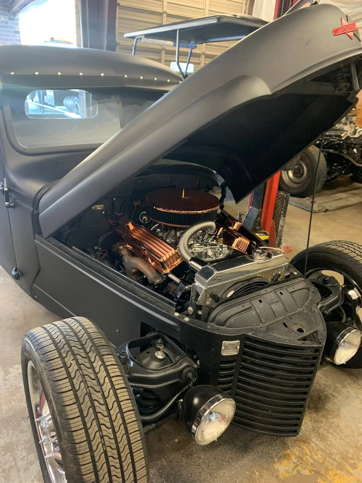 A black vintage hot rod car with its hood open, showing the engine with copper accents, in a garage with other vehicle parts and equipment in the background.