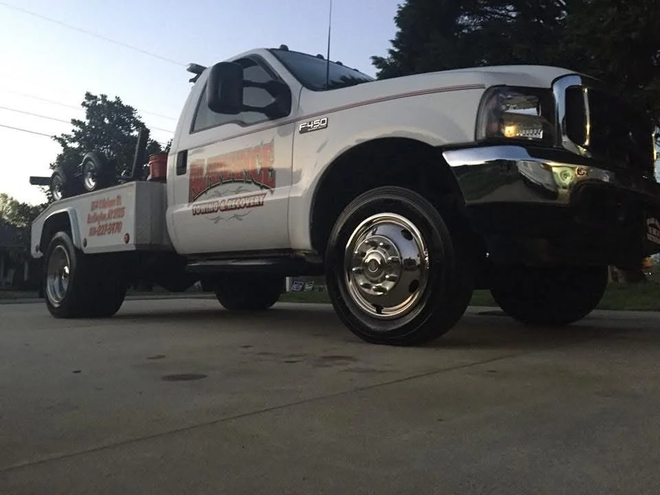 A white tow truck with a logo on the door, parked on a paved surface during dusk. A spare tire is mounted on the truck bed, and the vehicle has chrome hubcaps.