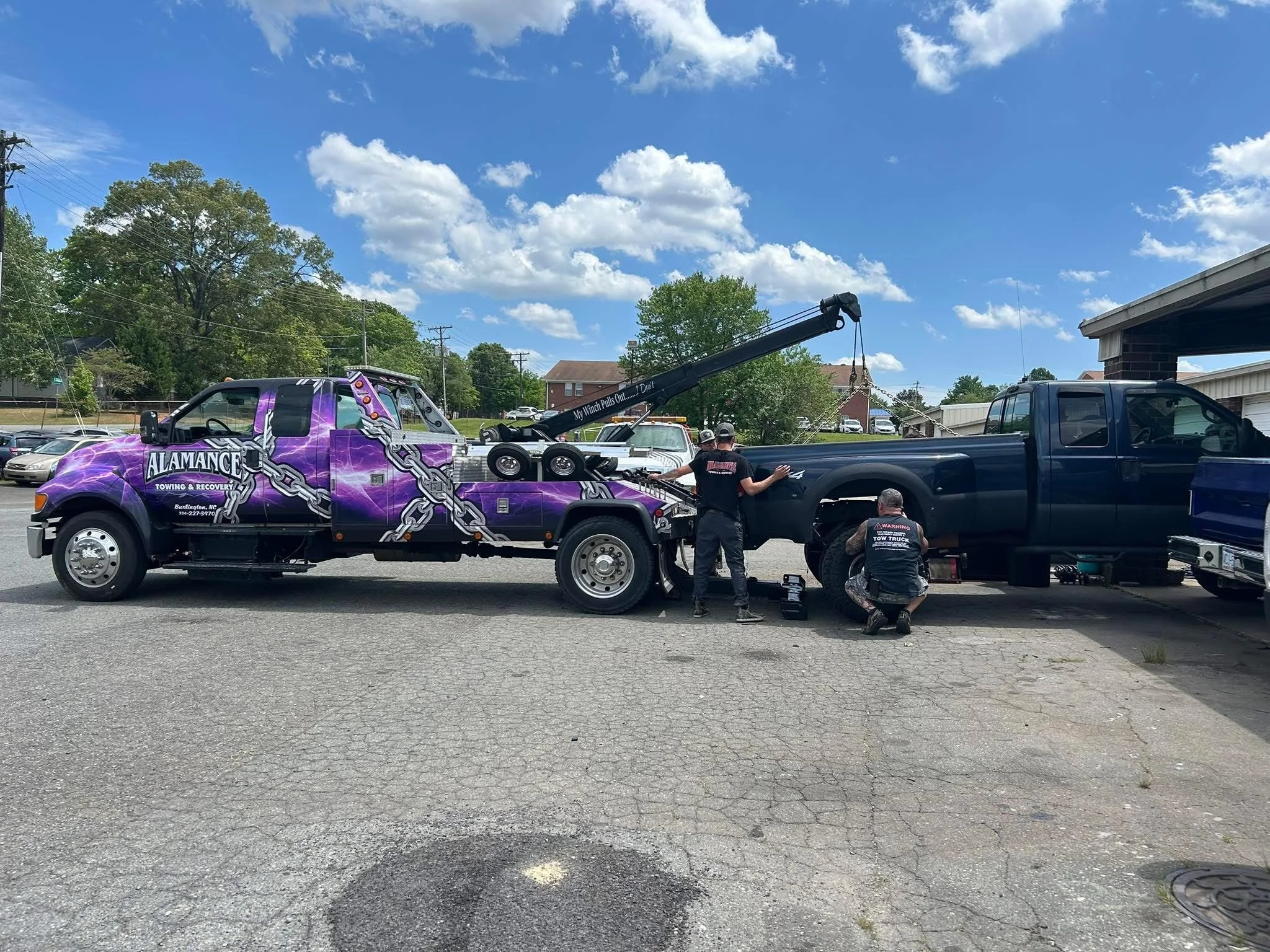 A tow truck painted purple with chains logos towing a black pickup truck in a parking lot on a sunny day, with four workers preparing the vehicle.