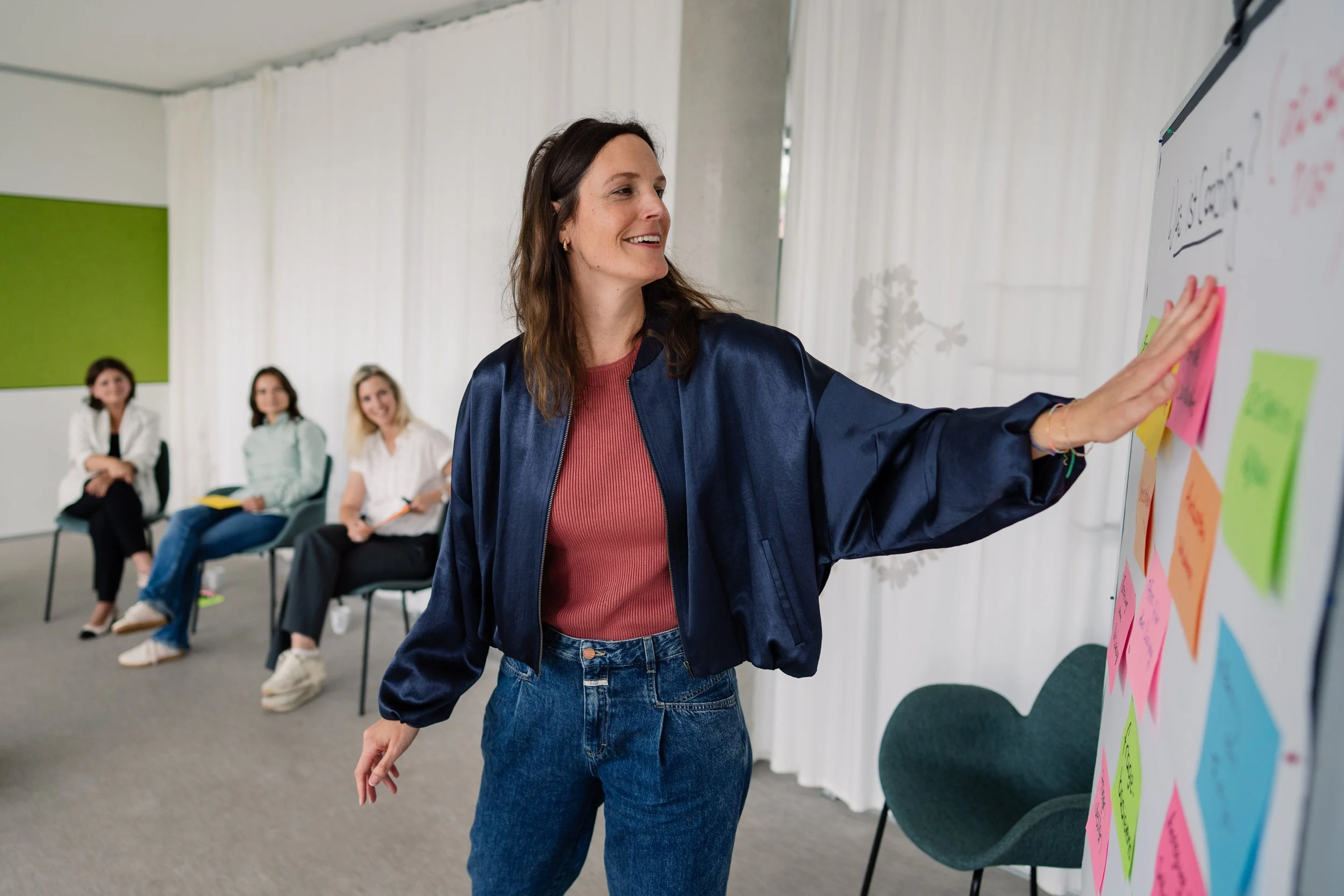 Eine Frau zeigt auf eine Whiteboard mit farbigen Haftnotizen während eines Workshops. Im Hintergrund sitzen drei Frauen, die beobachten und zuhören.