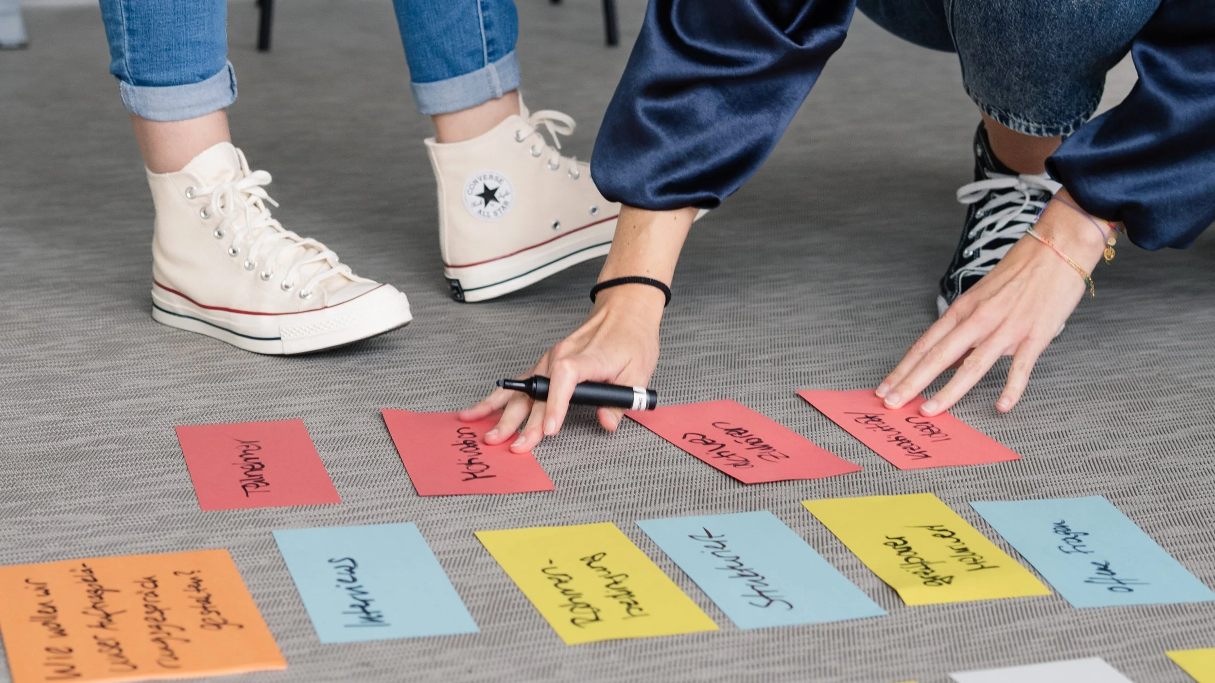 Person kniet auf Teppich und schreibt auf bunte Klebezettel, die auf dem Boden liegen, mit einem schwarzen Marker. Die Person trägt Jeans und Armbänder, während eine andere Person weißes Schuhwerk trägt, das Converse Logo zeigt.