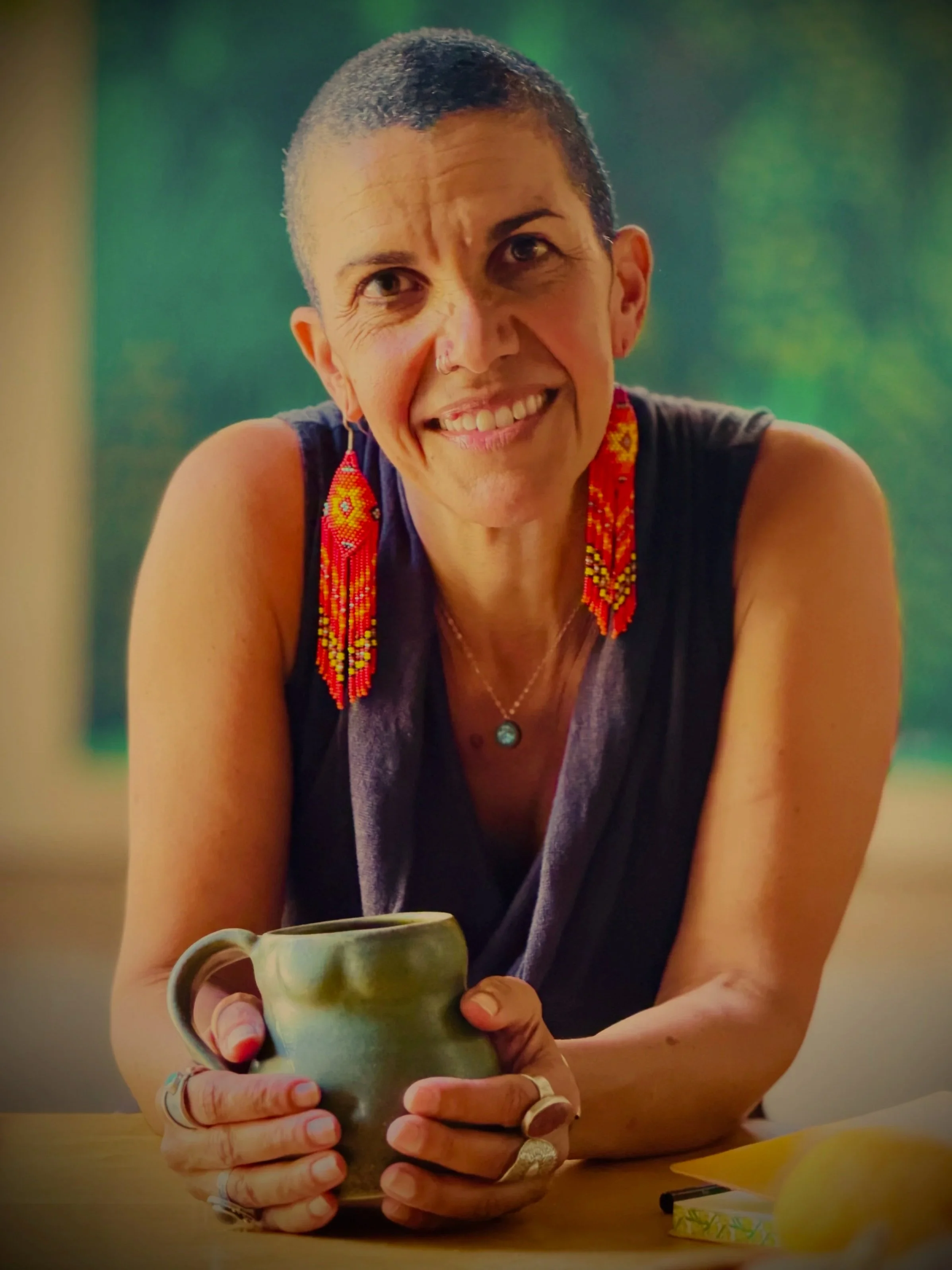 J. Miakoda Taylor wearing a brown tank top and red beaded earrings, smiles into the camera, warmly, while holding a mug of tea and wearing many rings