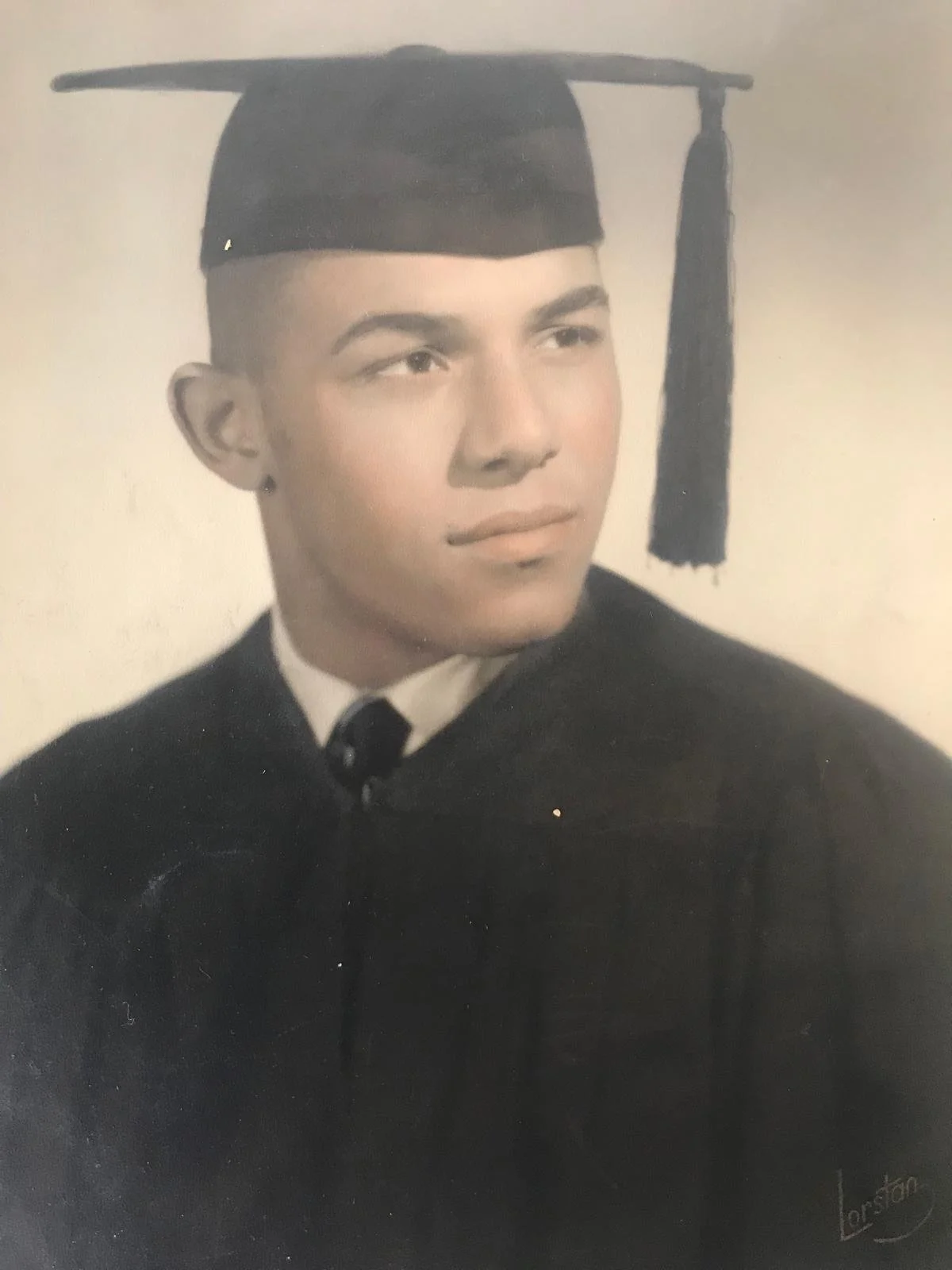 A sepia-toned photograph taken in the 1950s of Miakoda's father, as a young man, wearing a graduation cap and gown with a slight smile.