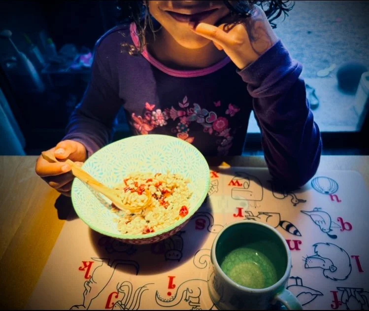 A 6-year-old girl with brown skin and brown curly hair, wearing a purple and pink flowered shirt, is sitting in front of a bowl with oatmeal with cranberries at the kitchen table. Her wooden spoon is poised to feed her. She has a smirk on her face.