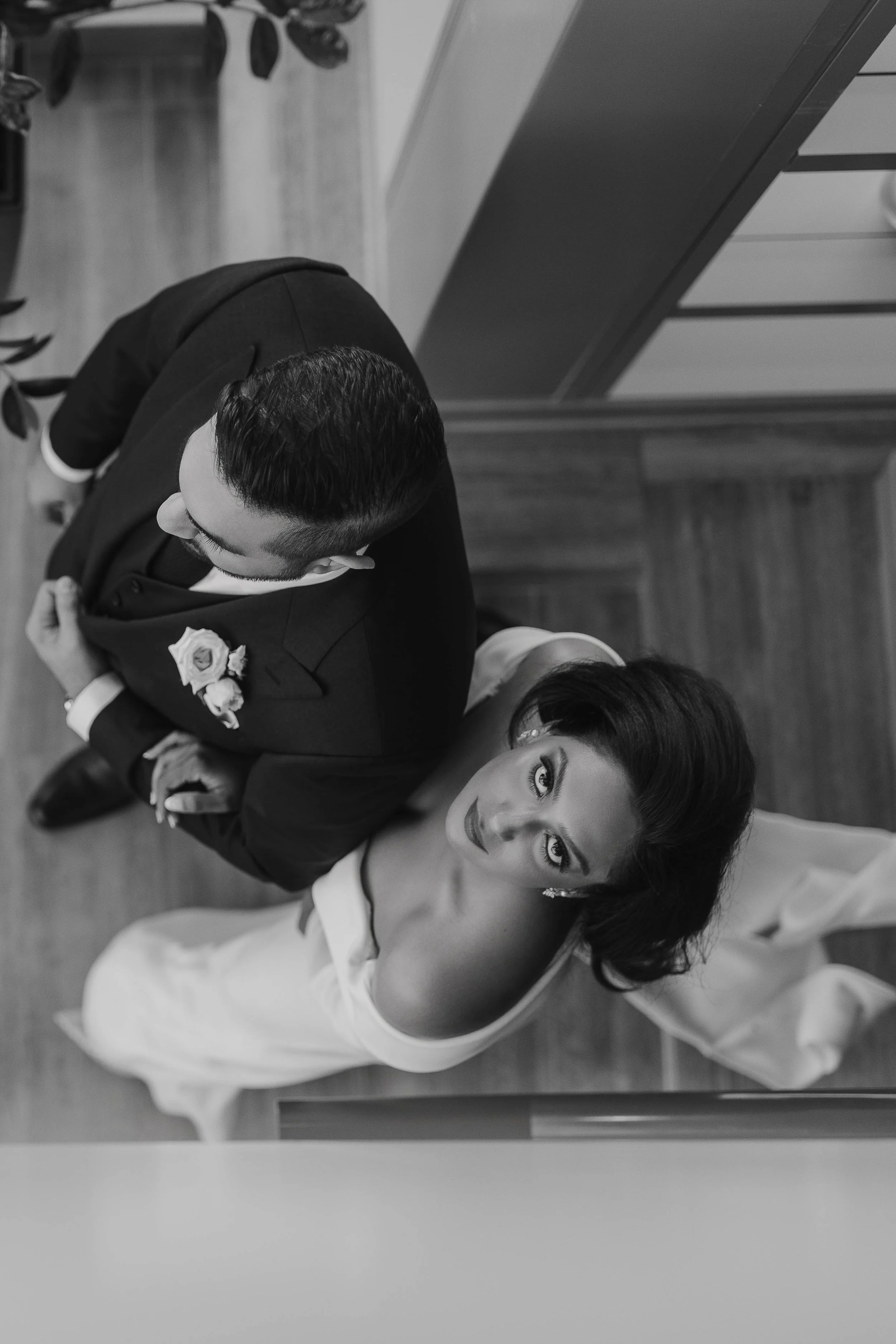 A black and white photo of a man and woman dressed in formal attire, possibly at a wedding, standing close together and looking up at the camera from an elevated angle.