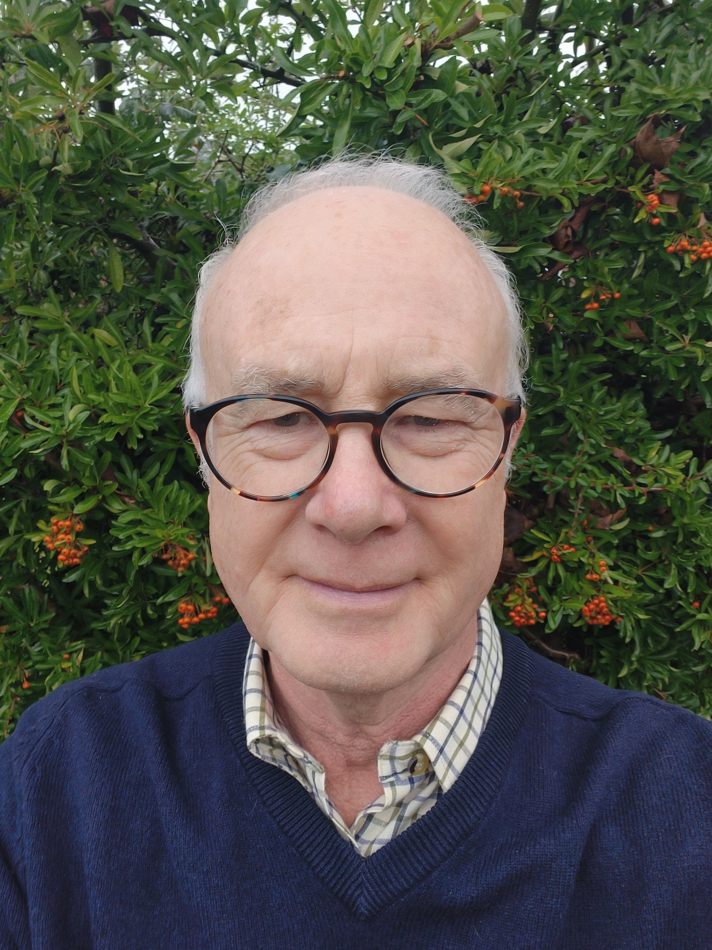A smiling older man with glasses, wearing a blue sweater over a plaid shirt, standing in front of green foliage with orange berries.