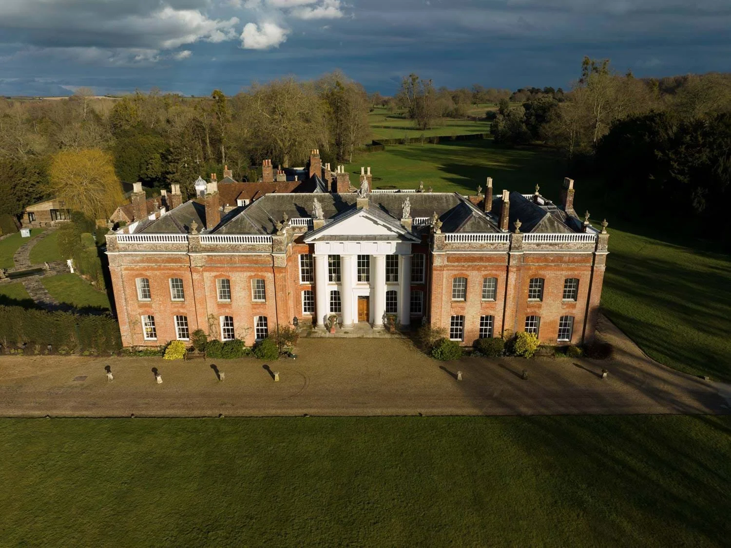 A large historic mansion with a red brick facade and white columns at the entrance, surrounded by manicured lawns and trees, under a cloudy sky.