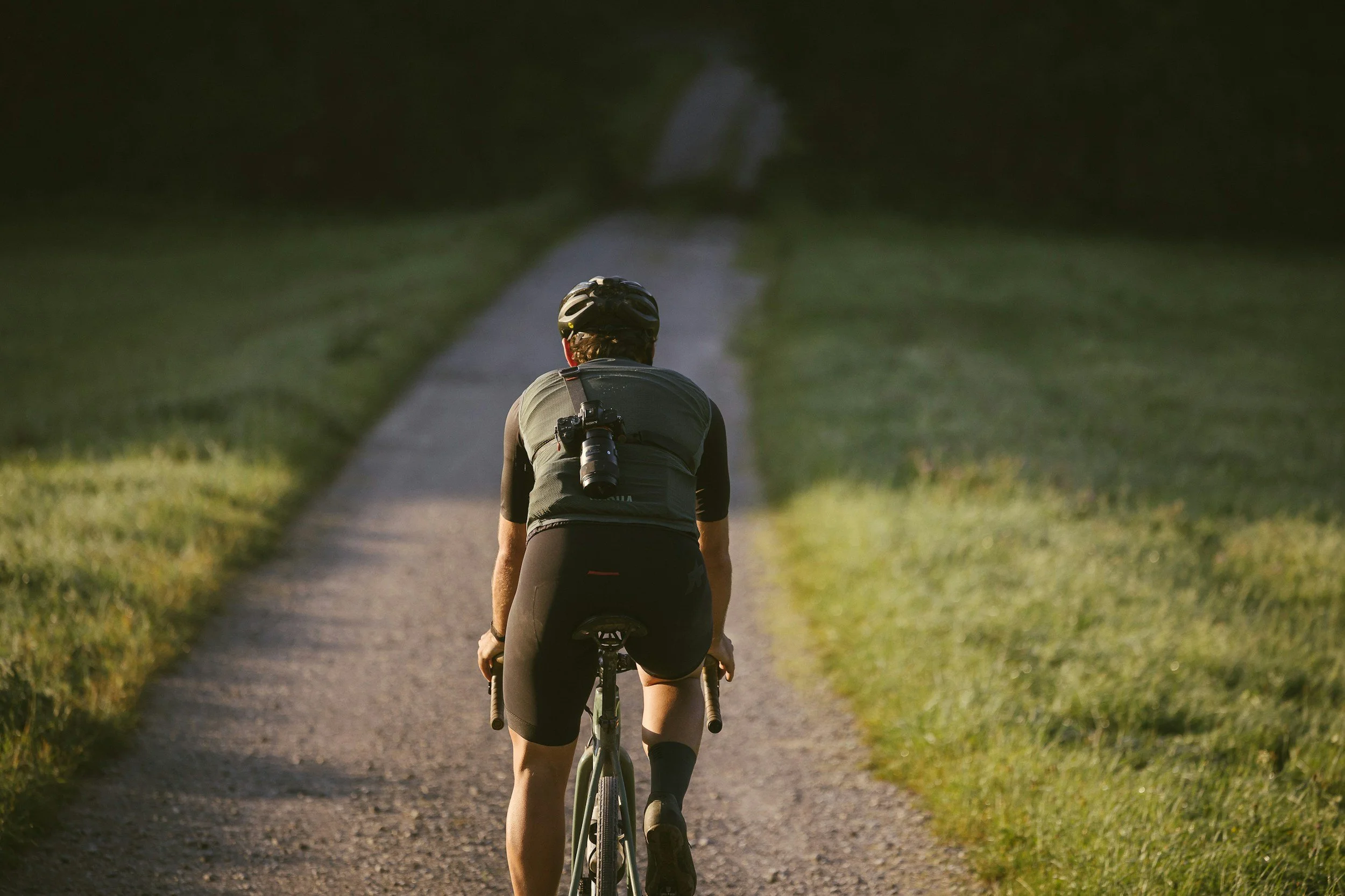 A person riding a bicycle on a gravel path through a grassy area, wearing a helmet and a camera hanging on their back.