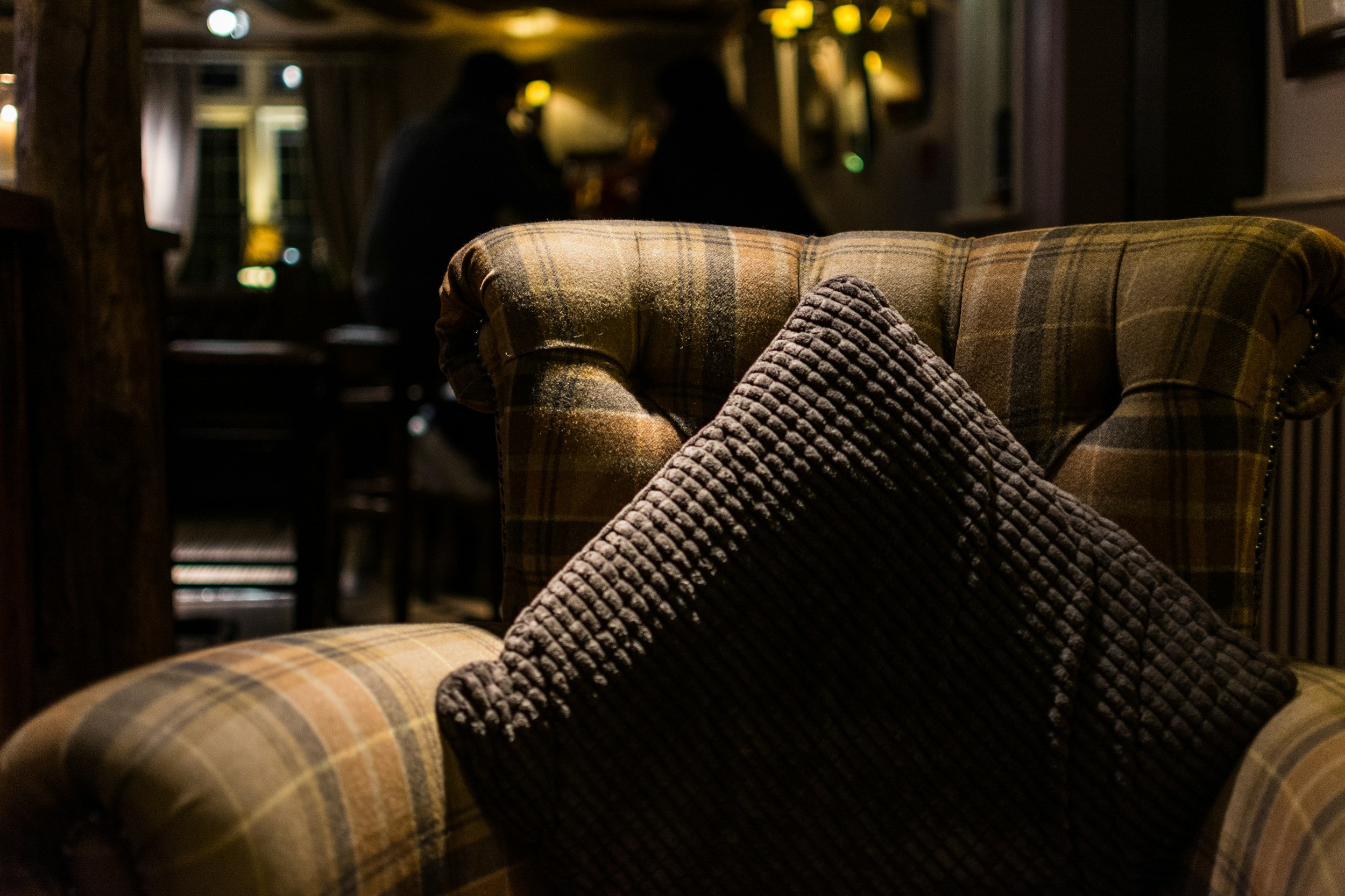 A plaid armchair with a dark textured cushion in a dimly lit room, with two silhouetted people talking in the background.