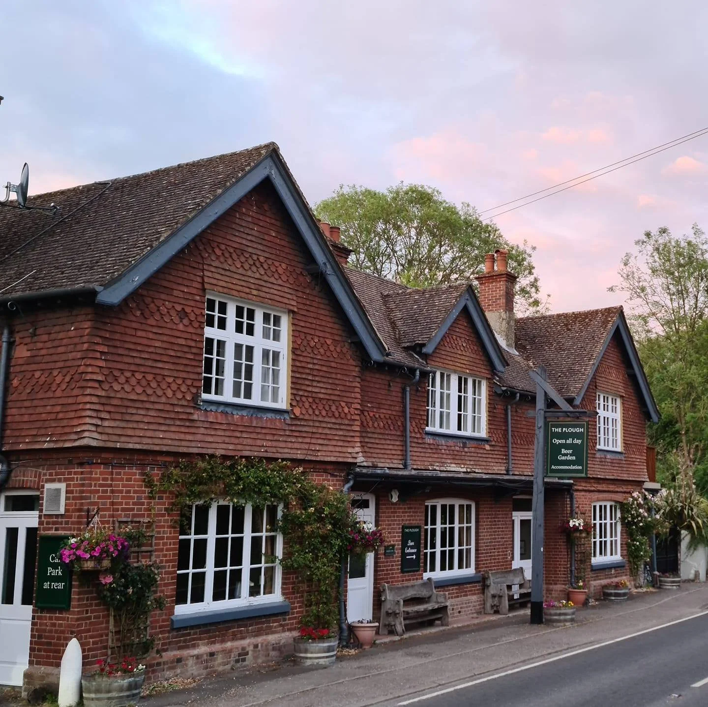 A traditional brick building with a sloped roof, white window frames, and hanging flower baskets. The building has signs indicating it is a pub called 'The Plough' and a car park at the rear. The sky is pink and purple, suggesting early evening or sunset.