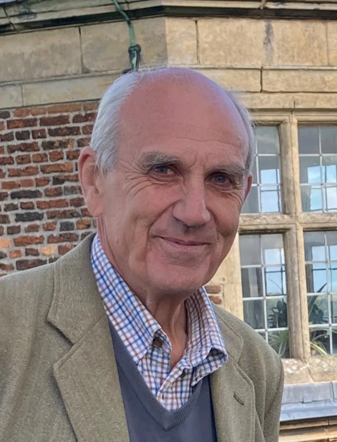 An elderly man with white hair, wearing a tan blazer, plaid shirt, and gray sweater, standing outside in front of a brick building with large windows.