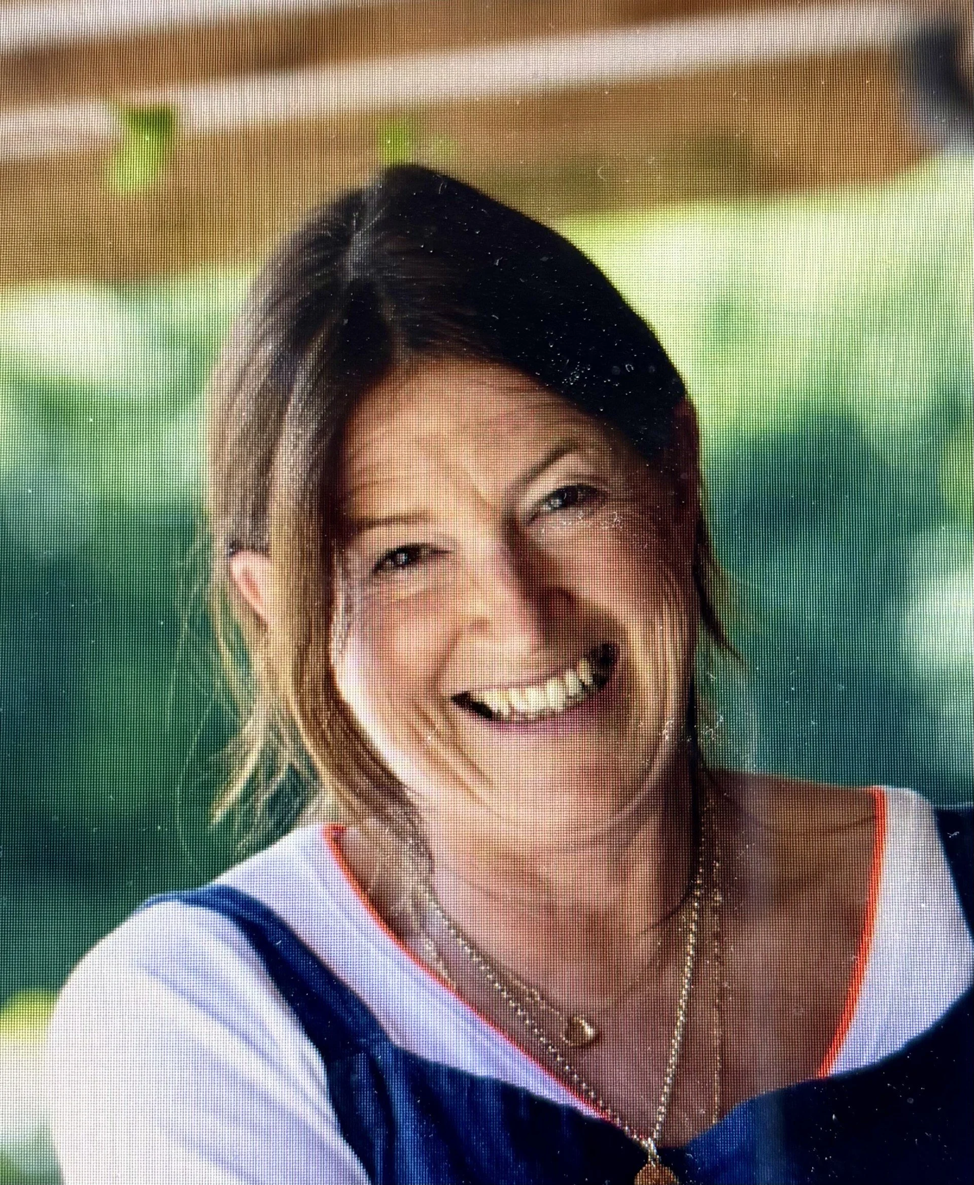 A woman smiling outdoors with a blurred green background. She has shoulder-length light brown hair and is wearing a dark top with white and orange accents, and layered necklaces.